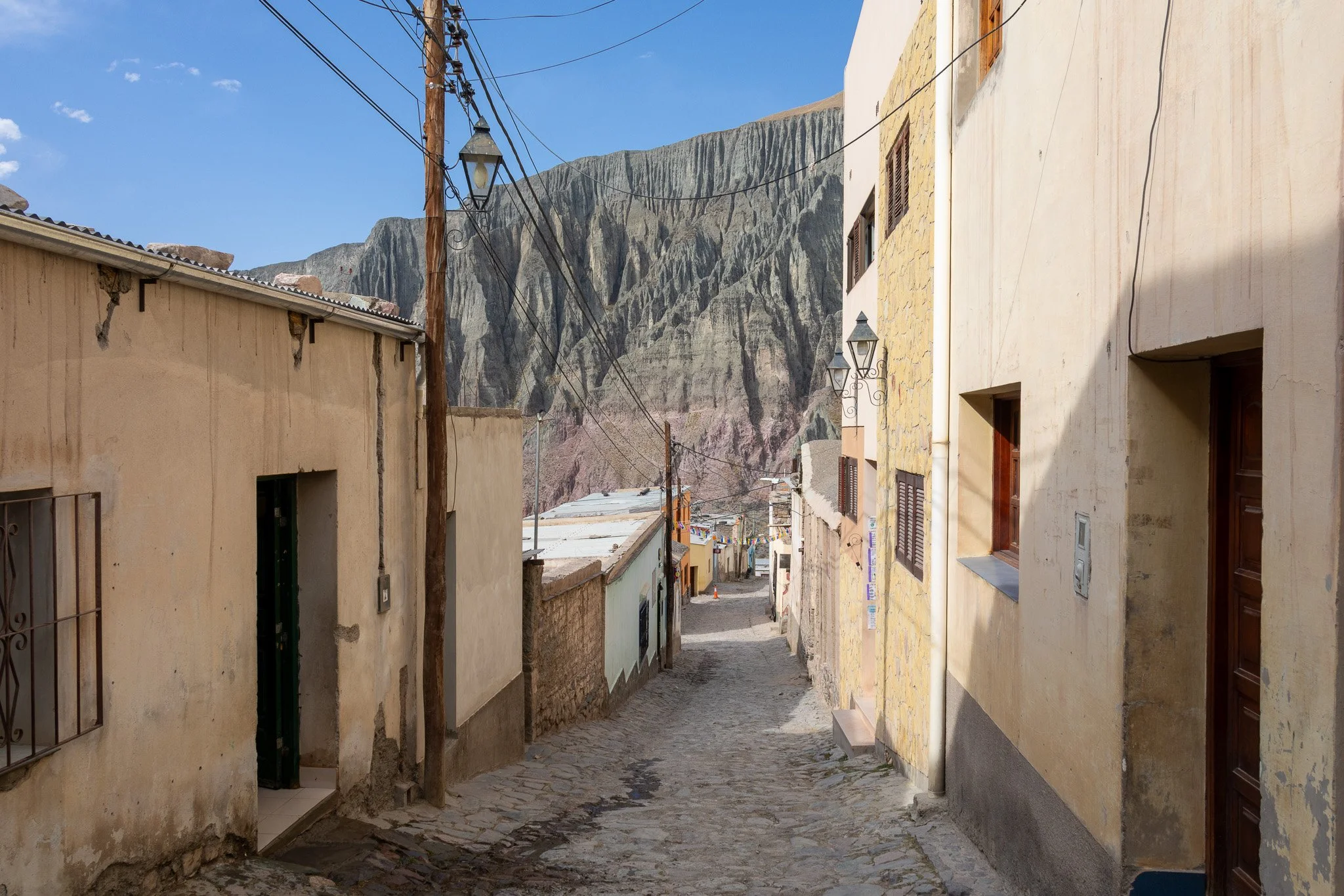 Cobblestone street in a small town with colorful buildings on either side, mountains in the background, and a clear blue sky.
