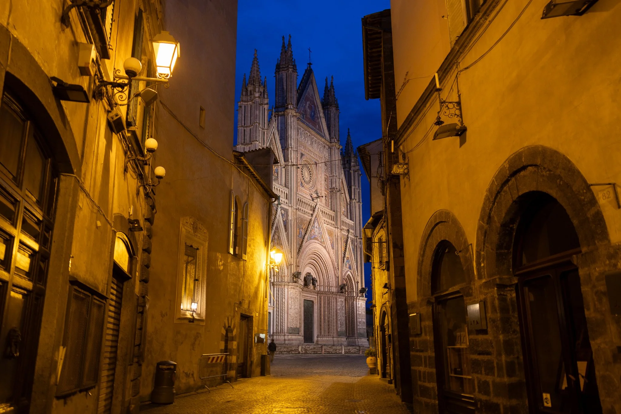Nighttime view of a narrow cobblestone street with an illuminated Gothic cathedral at the end, flanked by old buildings with yellow and brown walls, windows, and street lamps.