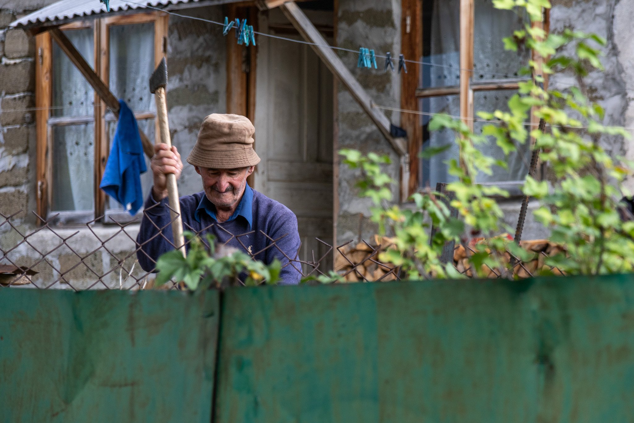 An elderly man wearing a hat and a blue sweater gardening behind a chain-link fence in front of a rustic house with boarded-up windows and laundry hanging on a clothesline.
