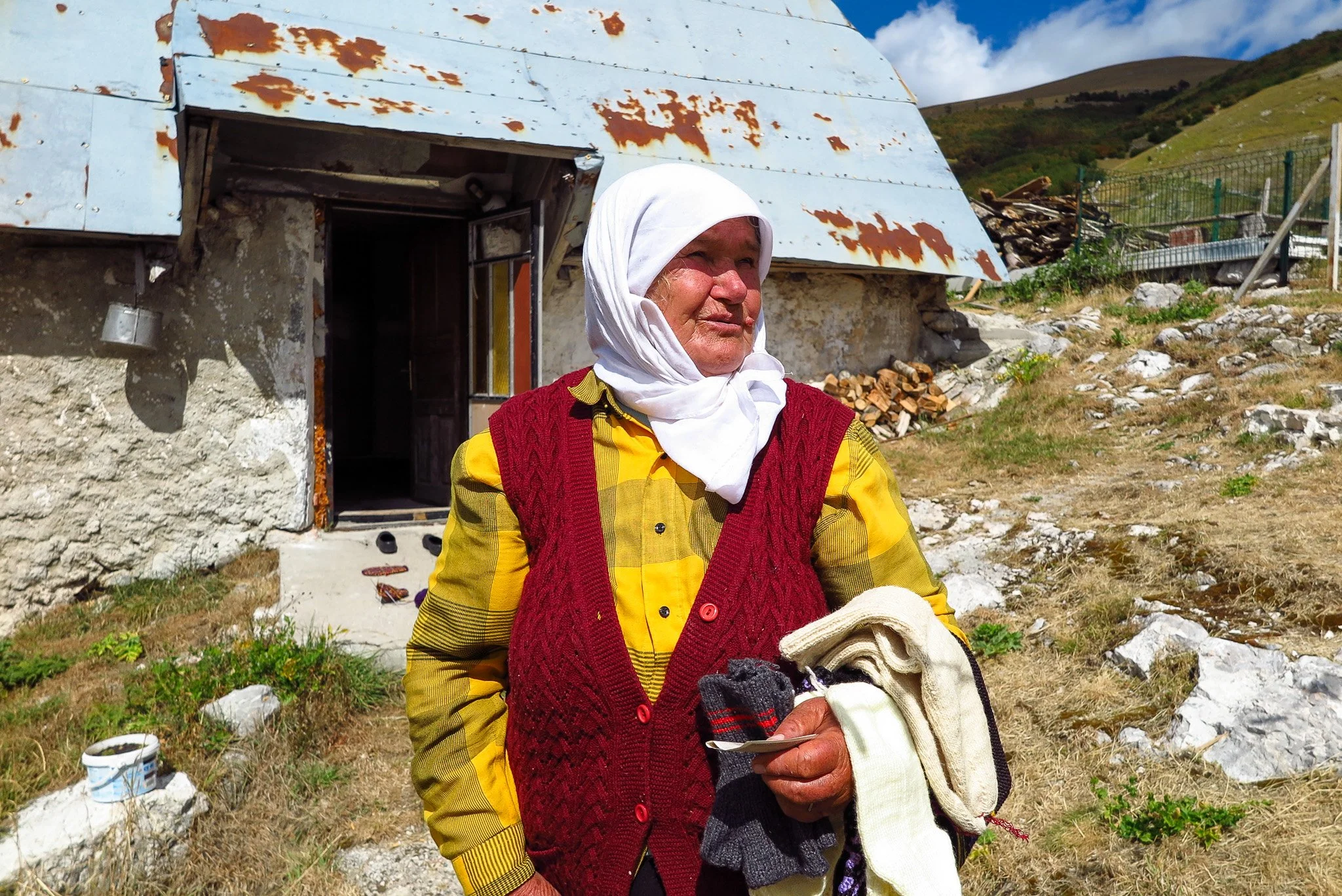 An elderly woman wearing a white headscarf, yellow shirt, and red vest stands outdoors near a rustic house with a rusty metal roof, with green hills in the background. She holds some knitted items and looks to the side with a gentle smile.