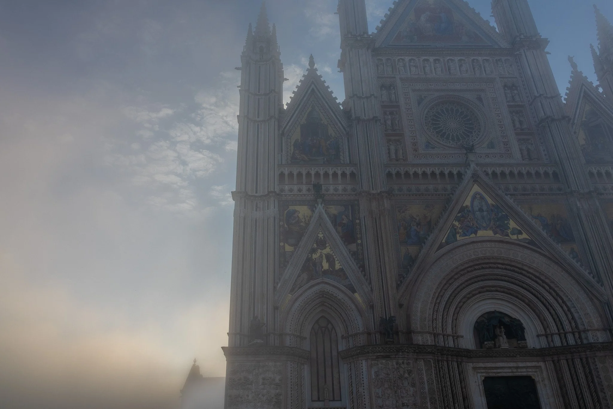 Part of a large, ornate cathedral with intricate stone carvings, stained glass windows, and a rose window, under a cloudy sky.