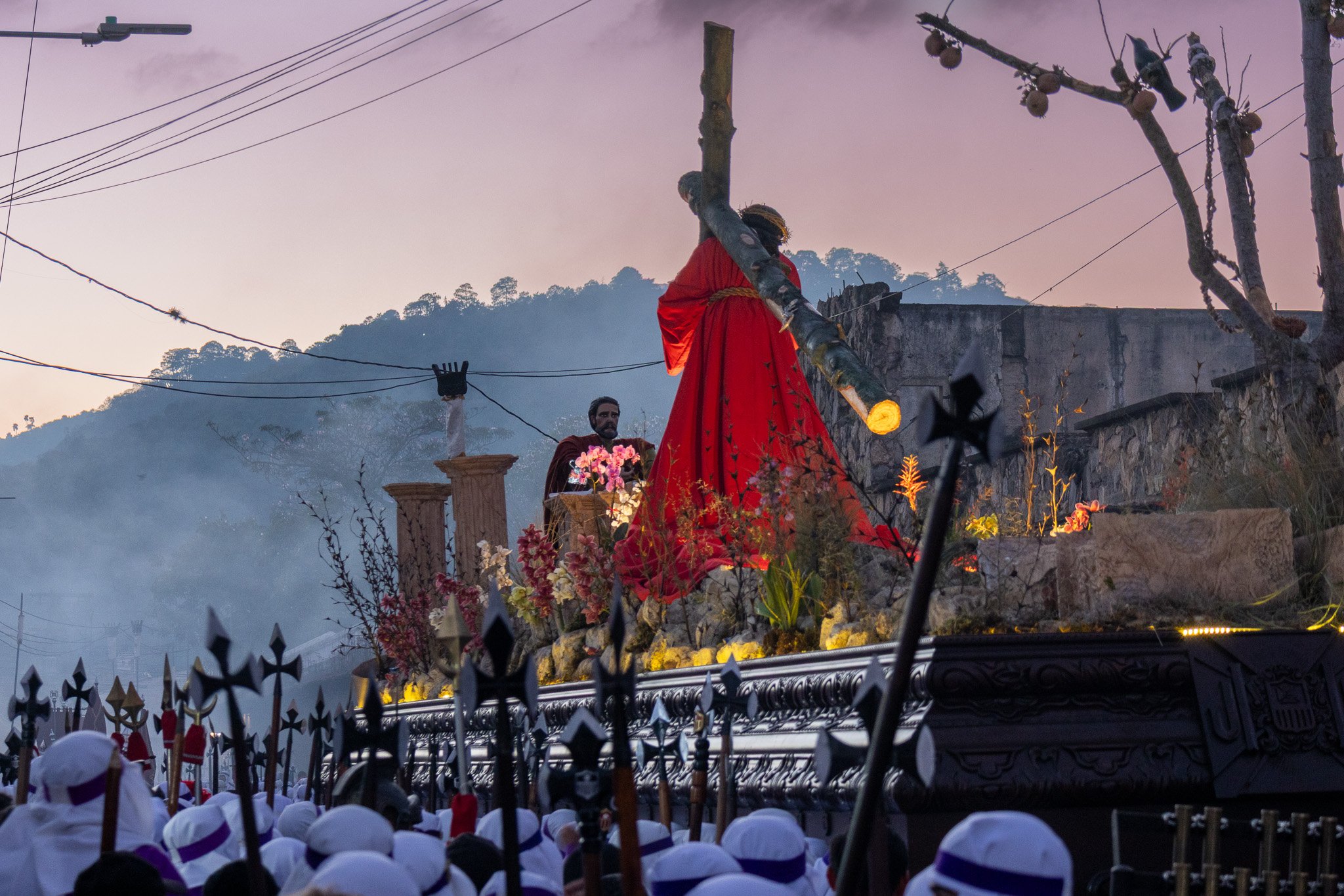 A religious procession with a statue of Jesus carrying a cross, dressed in a red robe, carried on a platform decorated with flowers. People in white hats are watching the procession, and a mountain with trees is visible in the background during sunse
