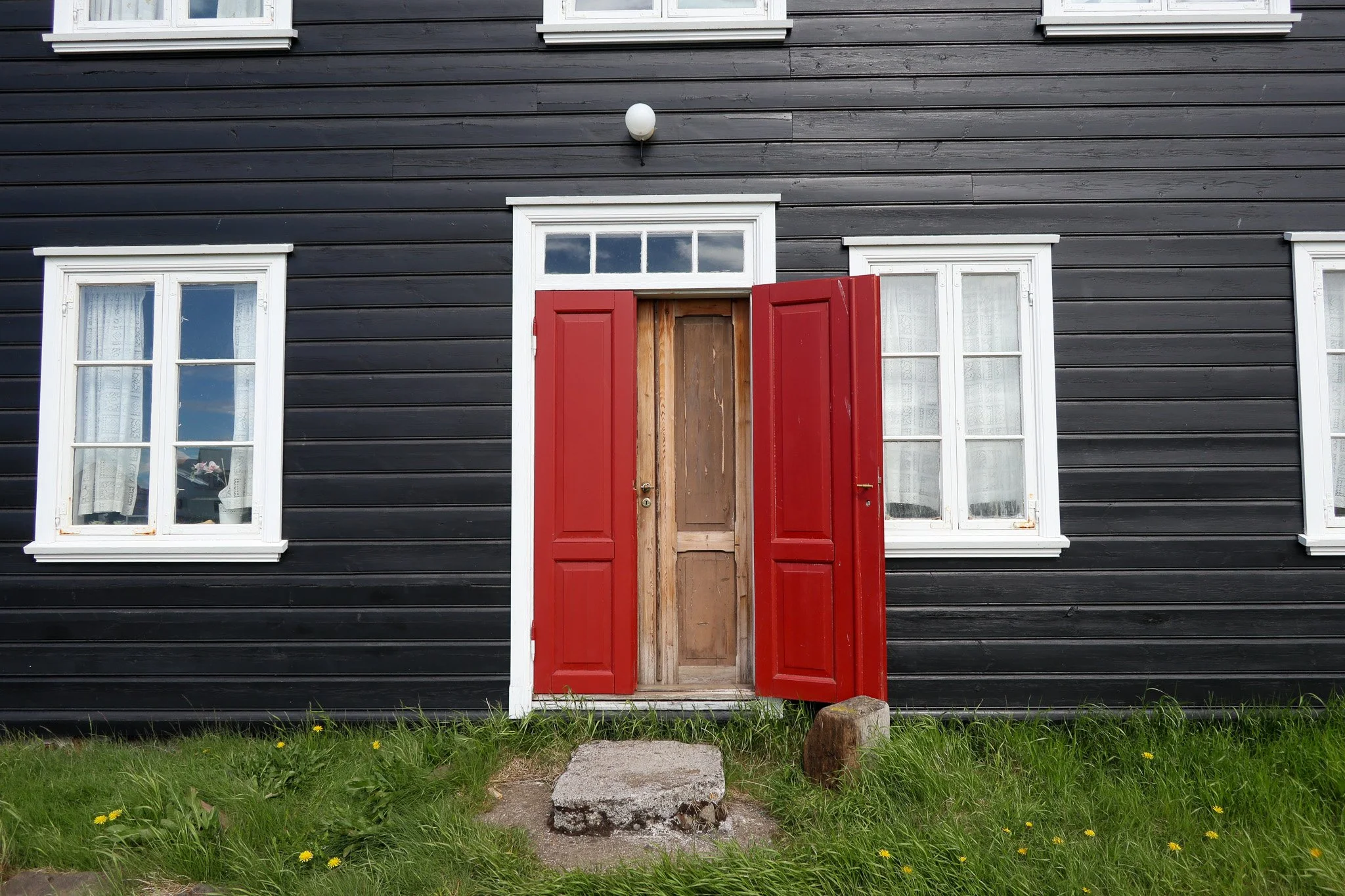A black wooden house with white-framed windows and red shutters, with an open wooden door, a stone step, and green grass with yellow flowers in front.