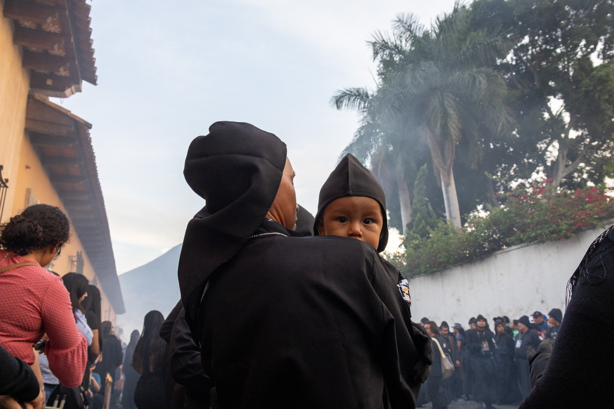 A woman holding a young child, both dressed in black hooded clothing, at a crowded outdoor gathering with trees and a building in the background.