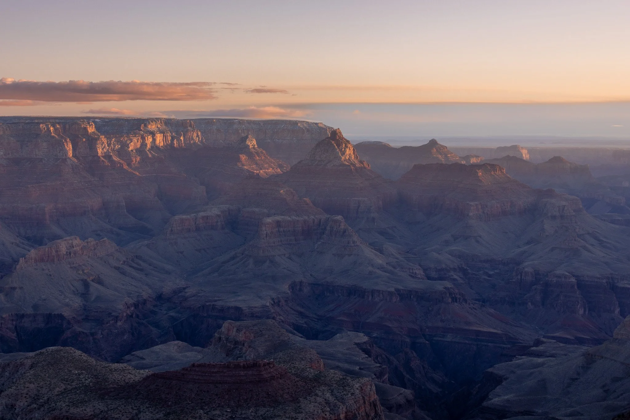 Wide view of the Grand Canyon at sunrise or sunset, showing layered rock formations and a partly cloudy sky.