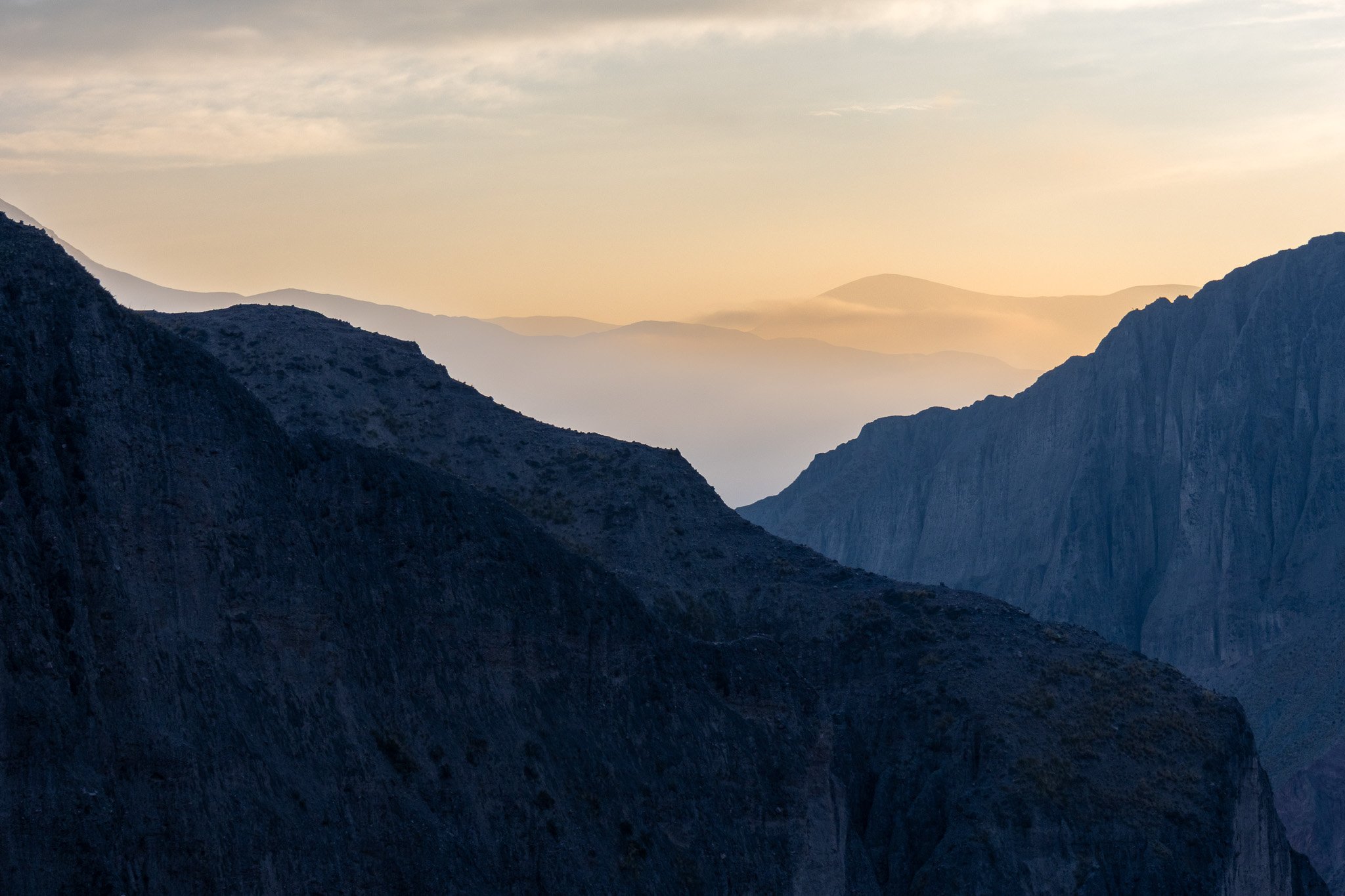 Mountain landscape with dark, rugged cliffs in the foreground and layers of mountains receding into the distance under a pale, cloudy sky at sunset or sunrise.