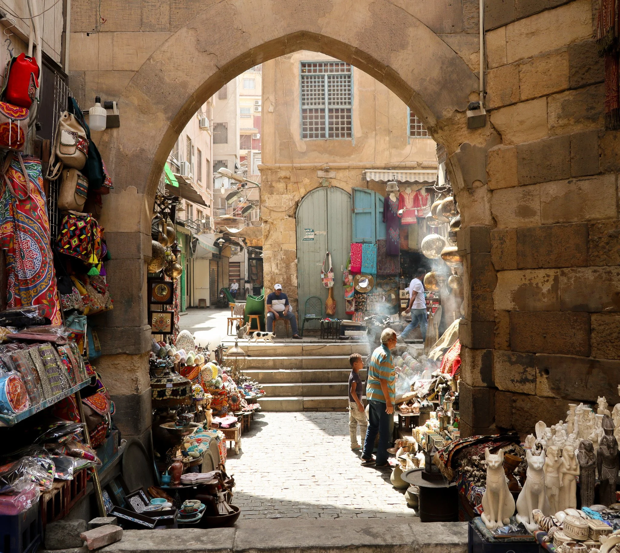 Street scene through an archway with vendors selling colorful bags, ceramics, and textiles, and people walking and sitting in an outdoor market