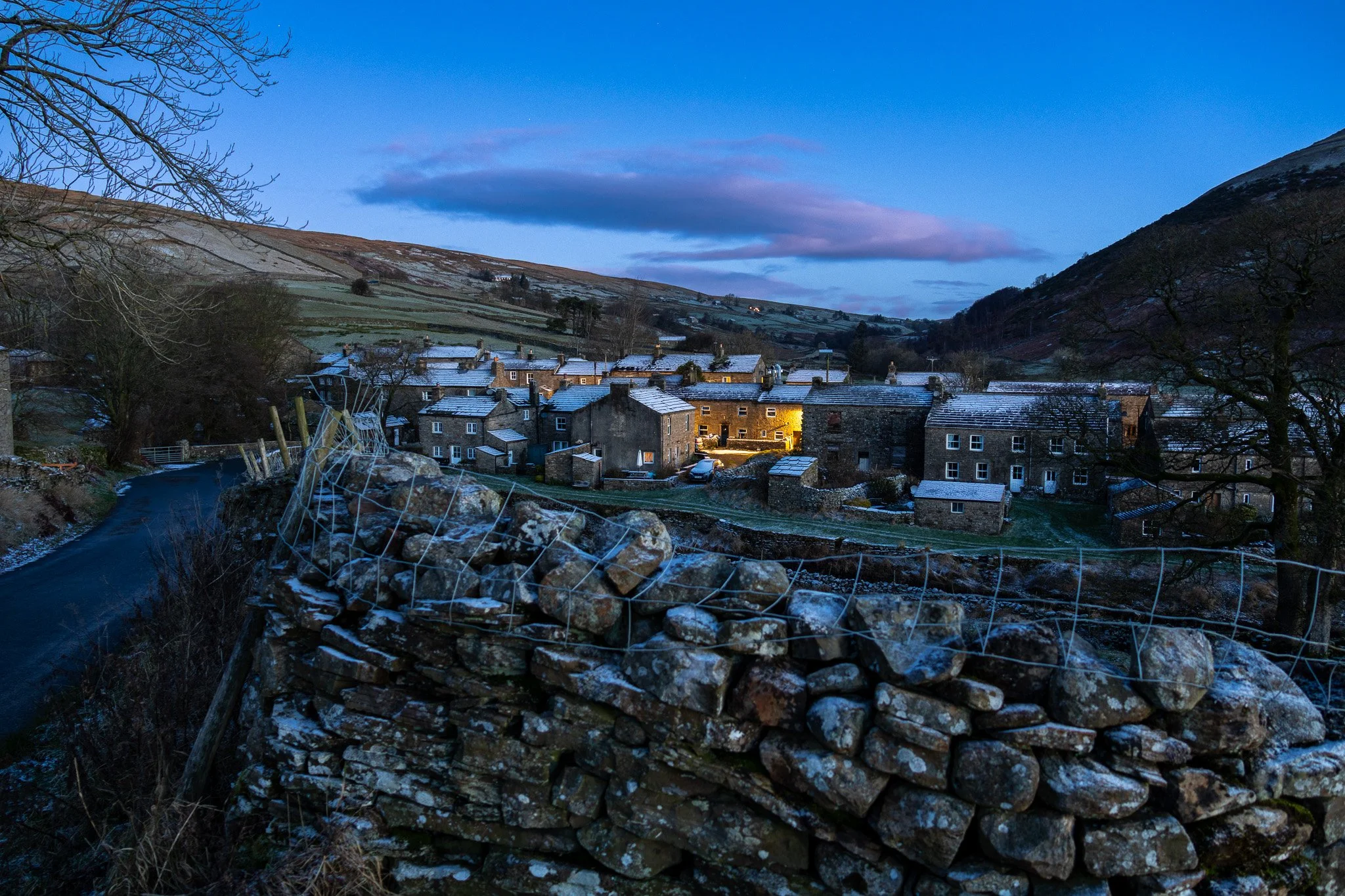 A quaint village nestled in a valley at dusk, with snow dusted rooftops and warm interior lights, surrounded by rolling hills and leafless trees, with a stone wall and a narrow road in the foreground.