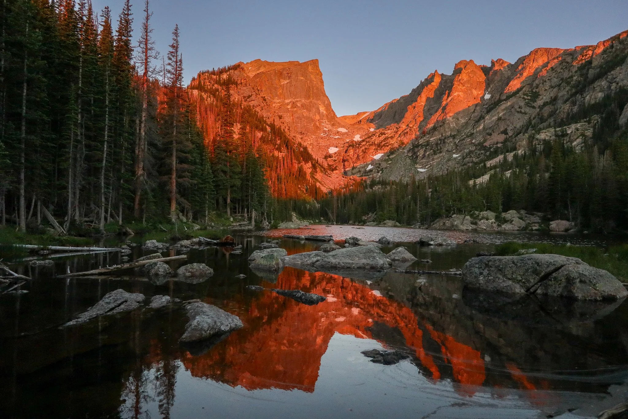 A mountain landscape at sunset with a peaceful river reflecting the orange-lit cliffs and pine trees.