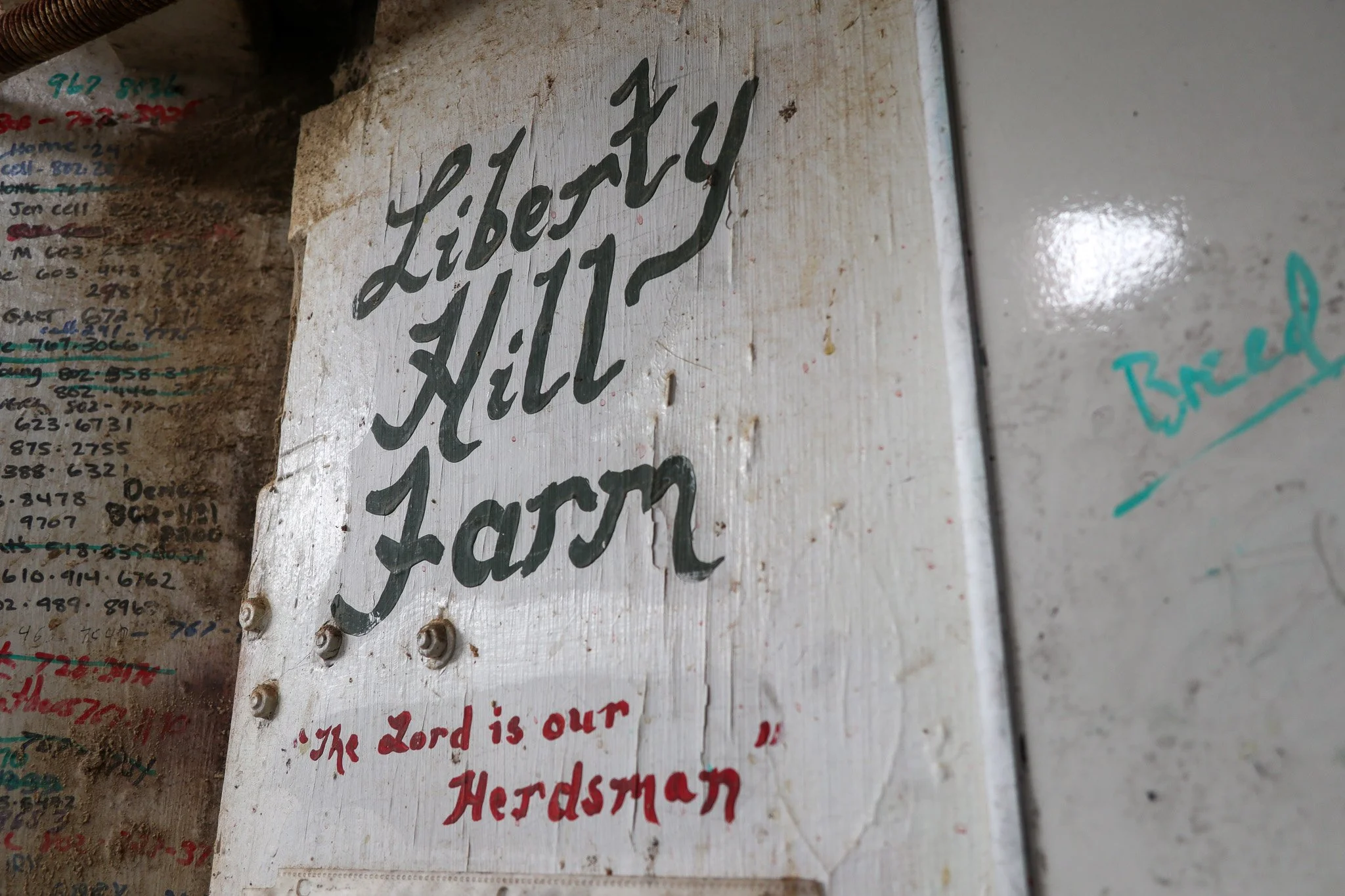 Hand-painted sign on a wooden surface with the words "Liberty Hill Farm" in large cursive letters and the phrase "The Lord is our Herdsman" in smaller red letters below.