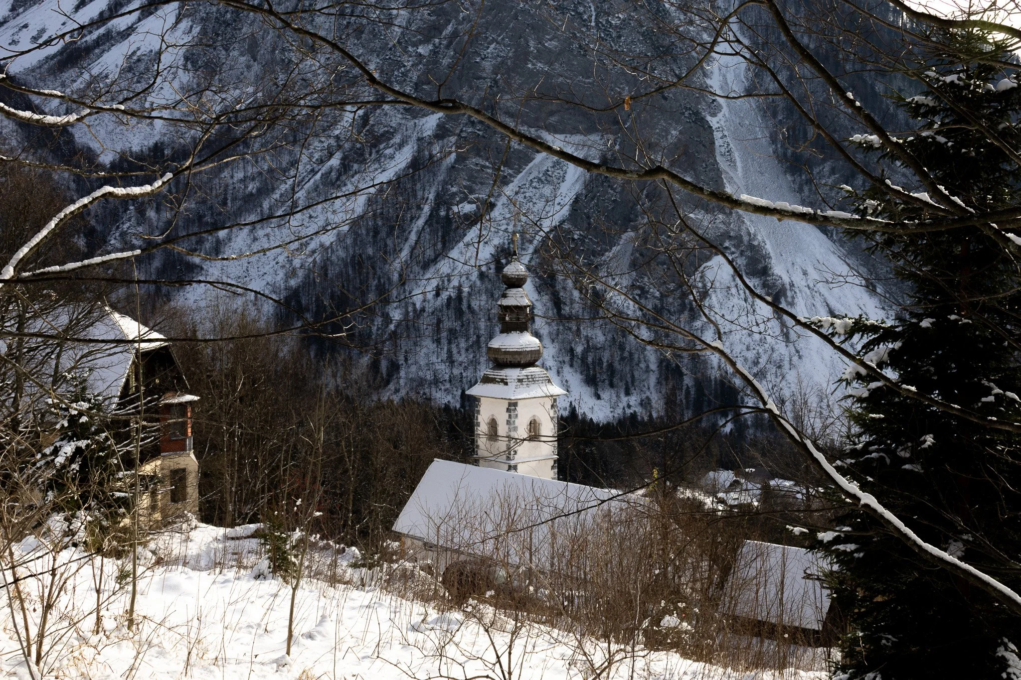 Snow-covered church with dark steeple amid snow-blanketed trees and mountain backdrop.