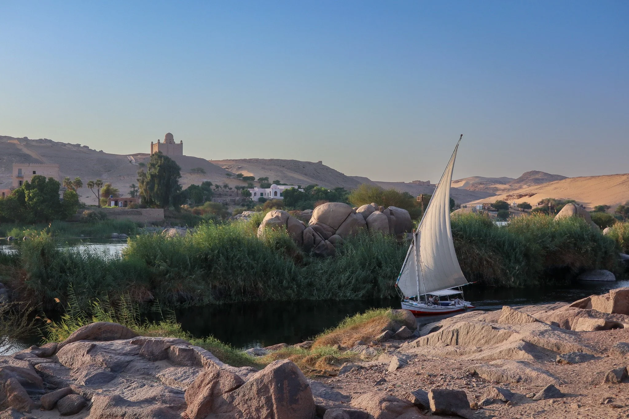 A sailboat on a river with lush green vegetation and rocks, surrounded by desert hills and a historic building in the distance at sunset.