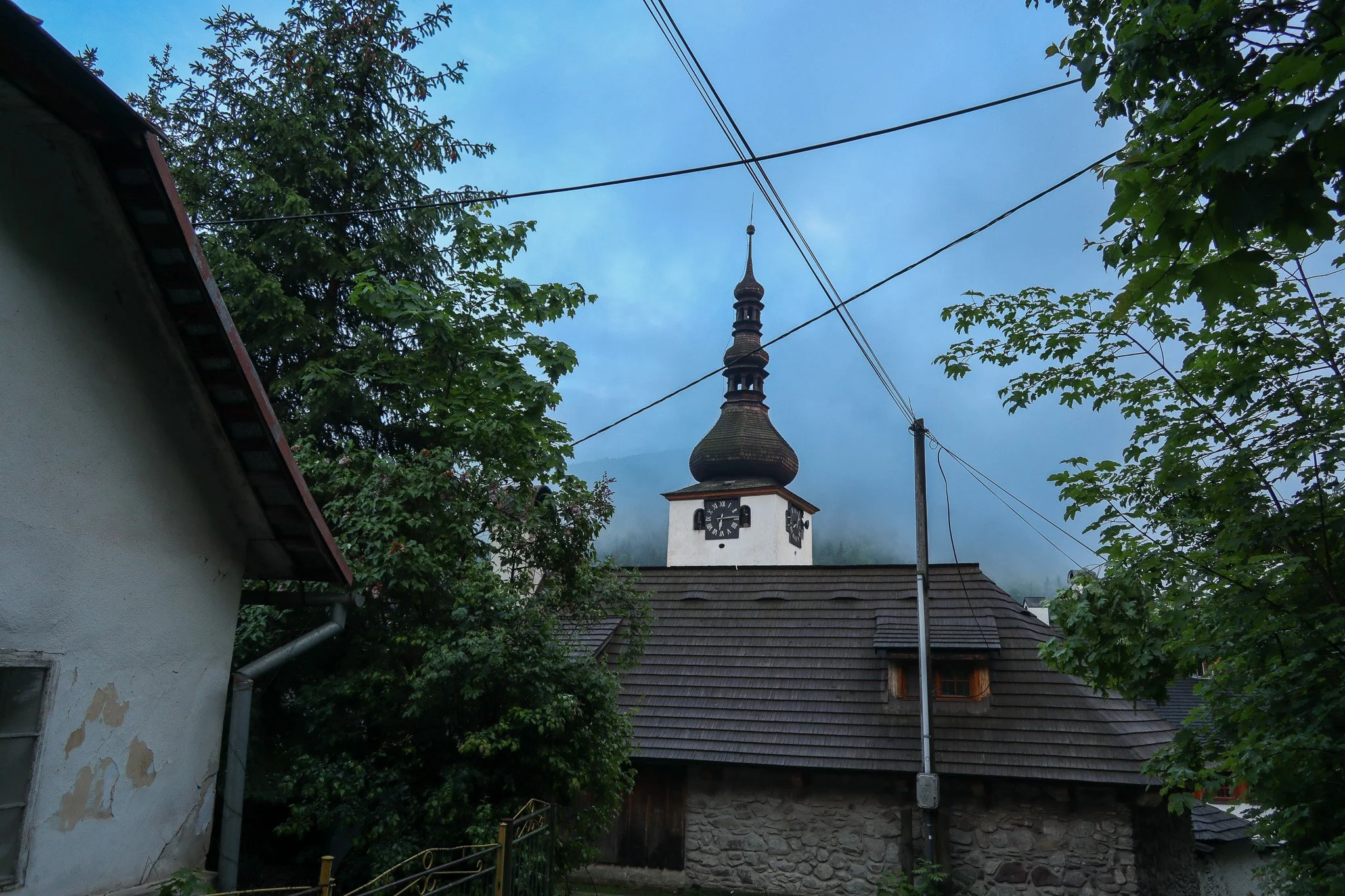 View of a church with a tall, ornate steeple, clock faces on the tower, surrounded by trees and electrical wires, under a partly cloudy sky.