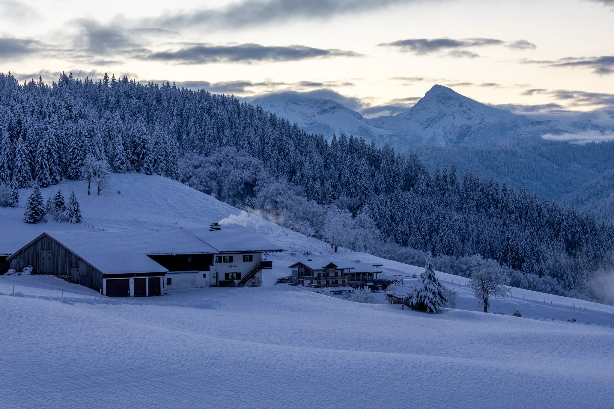 Snow-covered mountain landscape with trees, houses, and a forested mountain range in the background during winter.