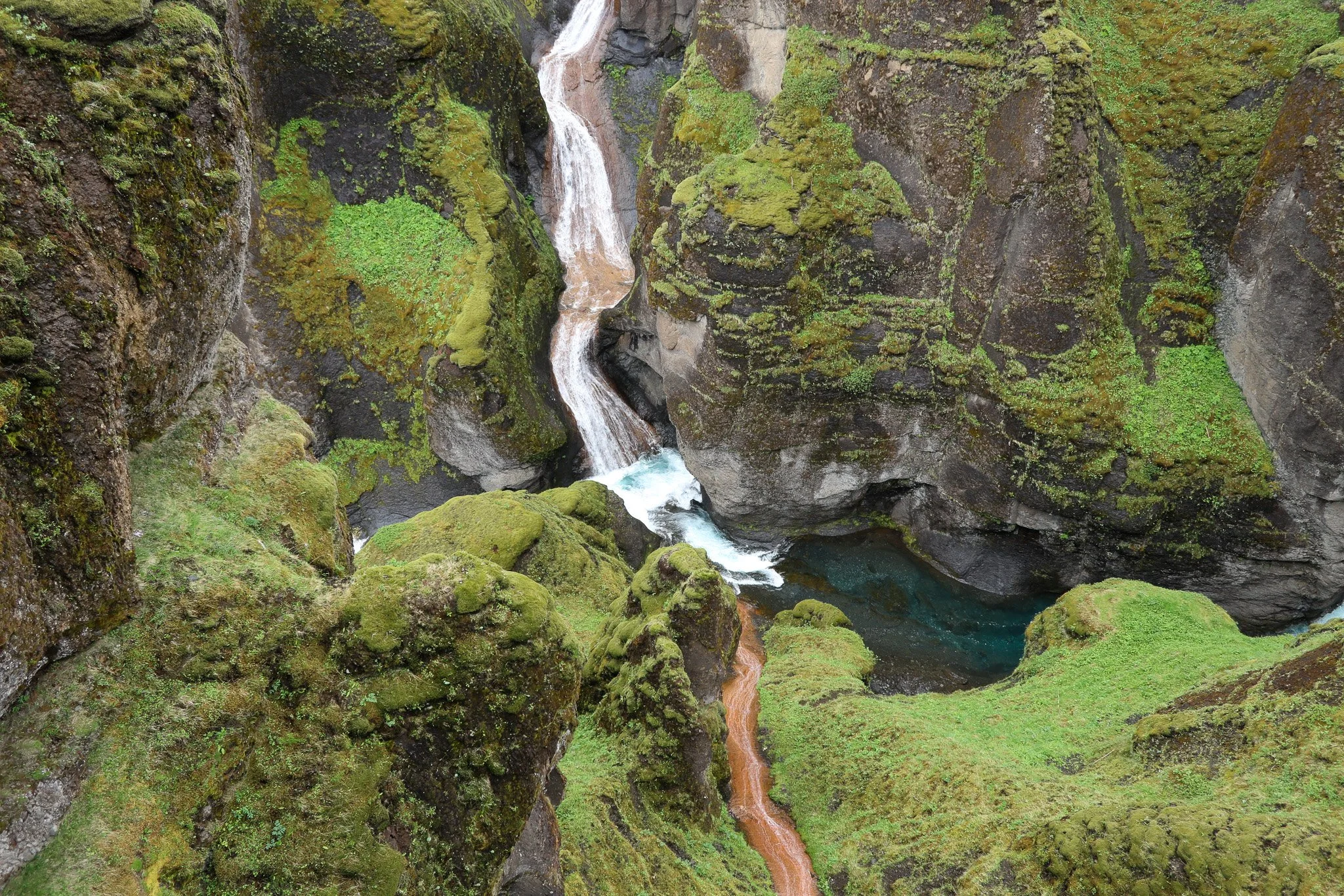 Aerial view of a narrow river flowing through a moss-covered canyon with waterfalls and lush green vegetation.