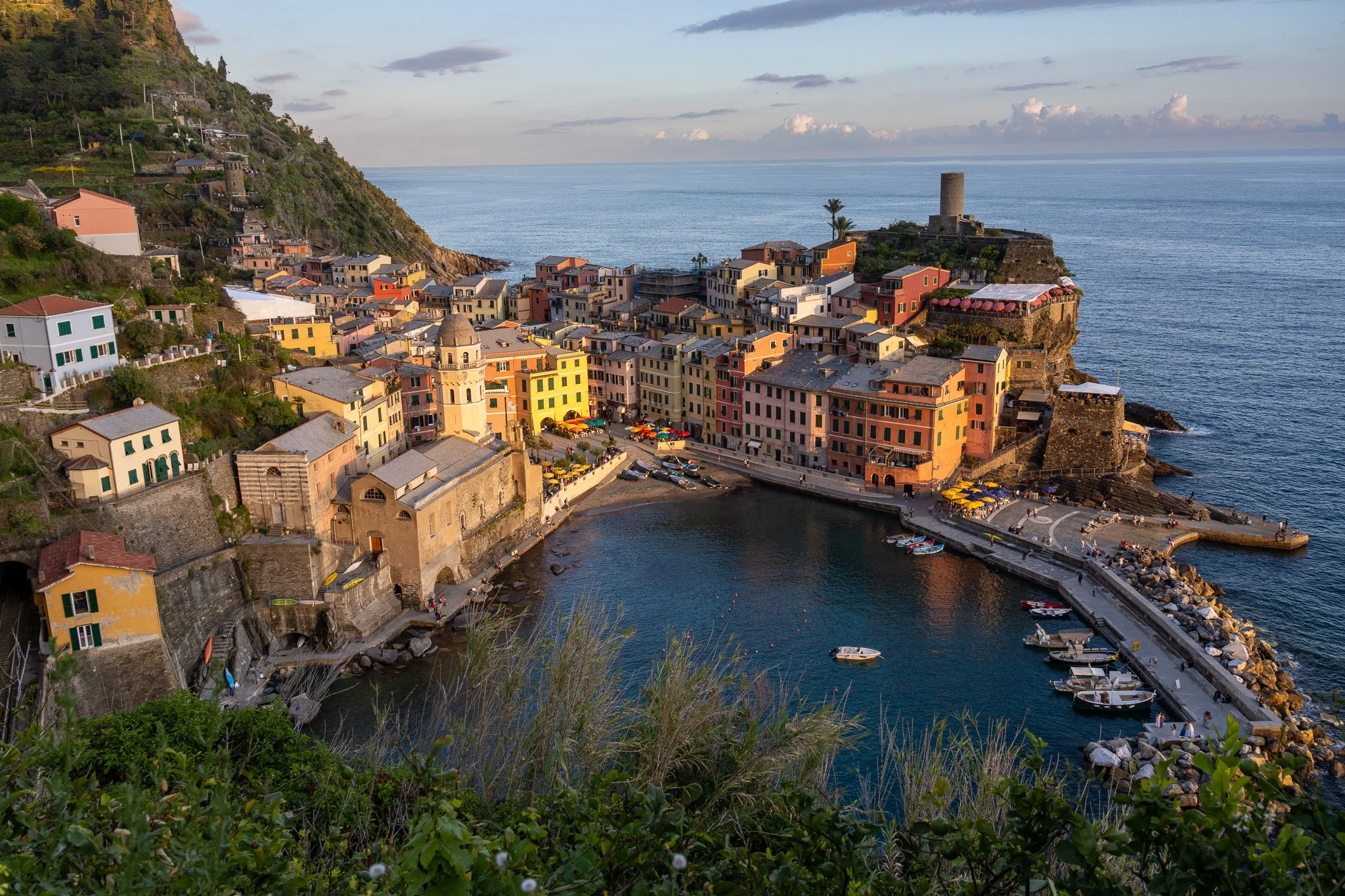 Colorful seaside village with pastel buildings on a hillside, a small harbor with boats, and a castle tower overlooking the ocean at sunset.
