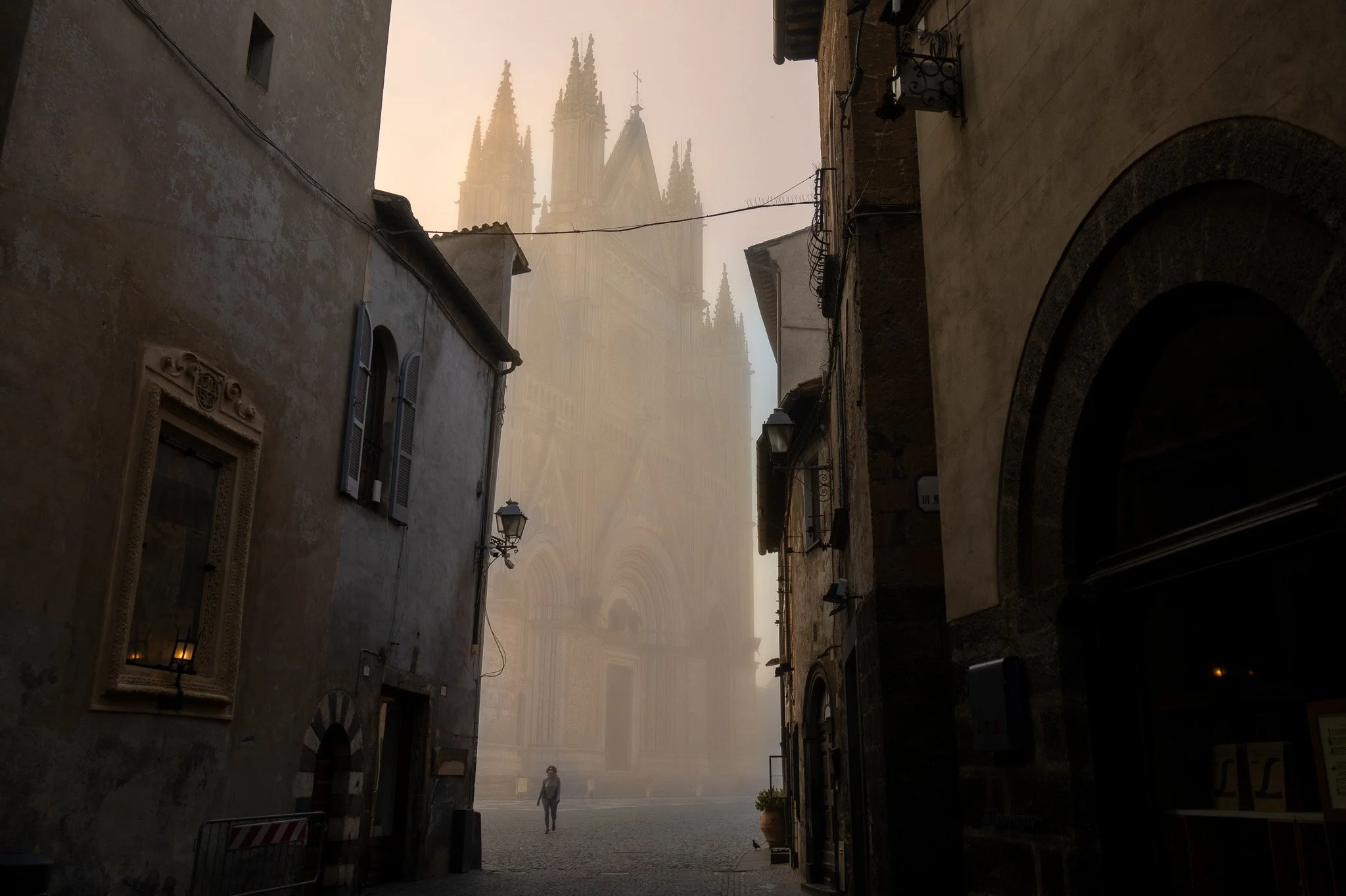 A narrow street in an old European city with tall, historic buildings, some with window shutters, leading towards a large cathedral in the background with tall spires, partially obscured by fog.