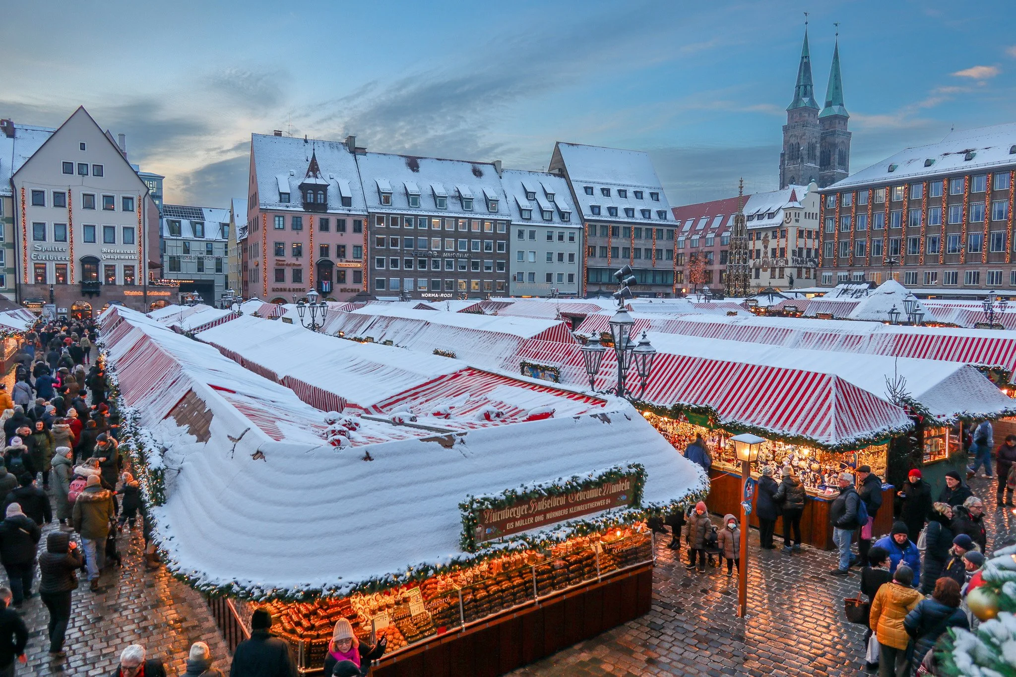 A lively Christmas market in a European city square during winter, with snow-covered stalls decorated with lights, and people shopping and walking around. Historic buildings with snow on their roofs and a church with two tall spires are visible in th