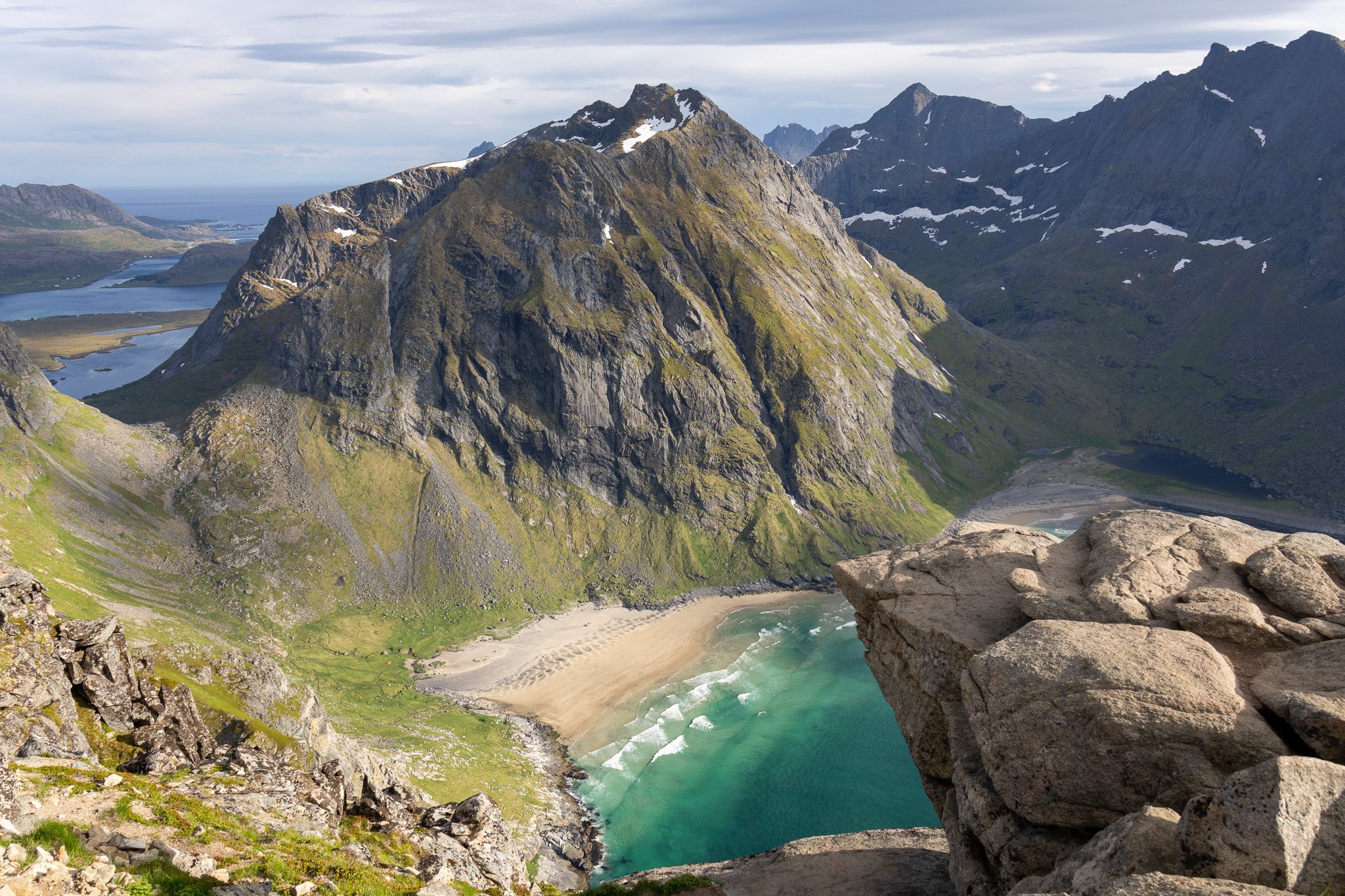 A scenic view of a coastal landscape with large moss-covered mountains, a sandy beach, and turquoise waters, viewed from a rocky vantage point.