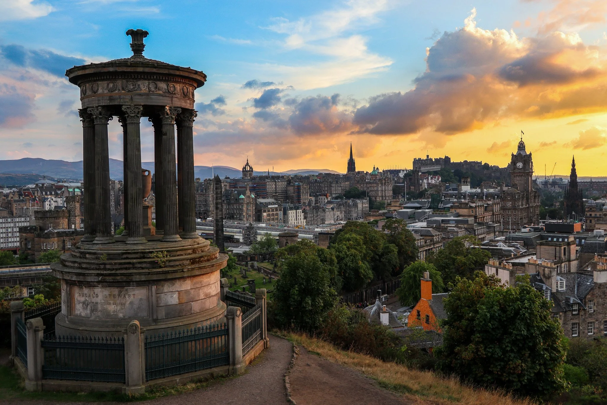 Historical monument with columns overlooking a cityscape at sunset, featuring old buildings, church spires, and trees.