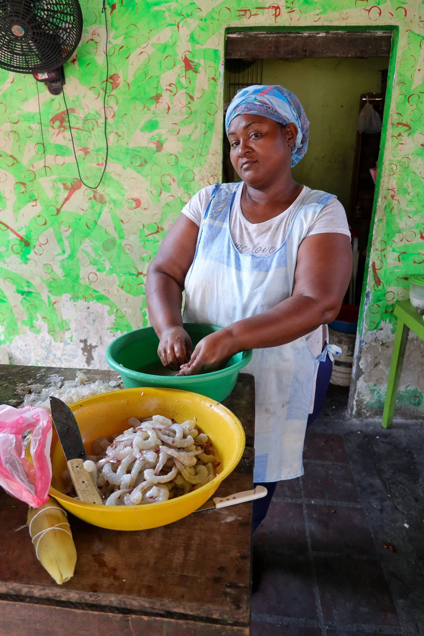A woman wearing a headscarf and apron washing shrimp in a green basin in a kitchen with colorful, patterned walls. There is a basket of peeled shrimp, a knife, and a bunch of plantains on the table.