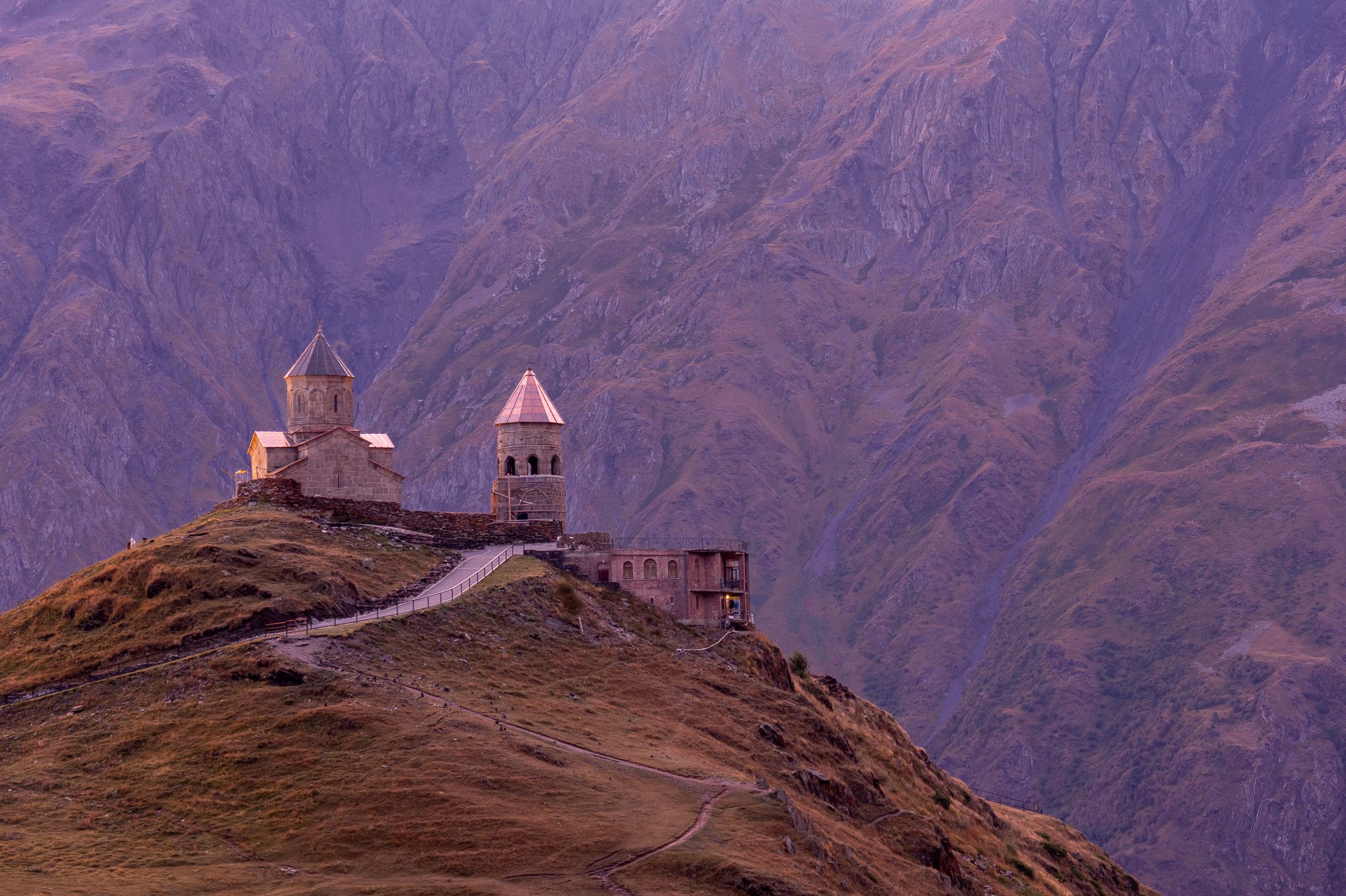 A small stone church with two towers located on a hilltop in a mountainous landscape with rugged peaks in the background.