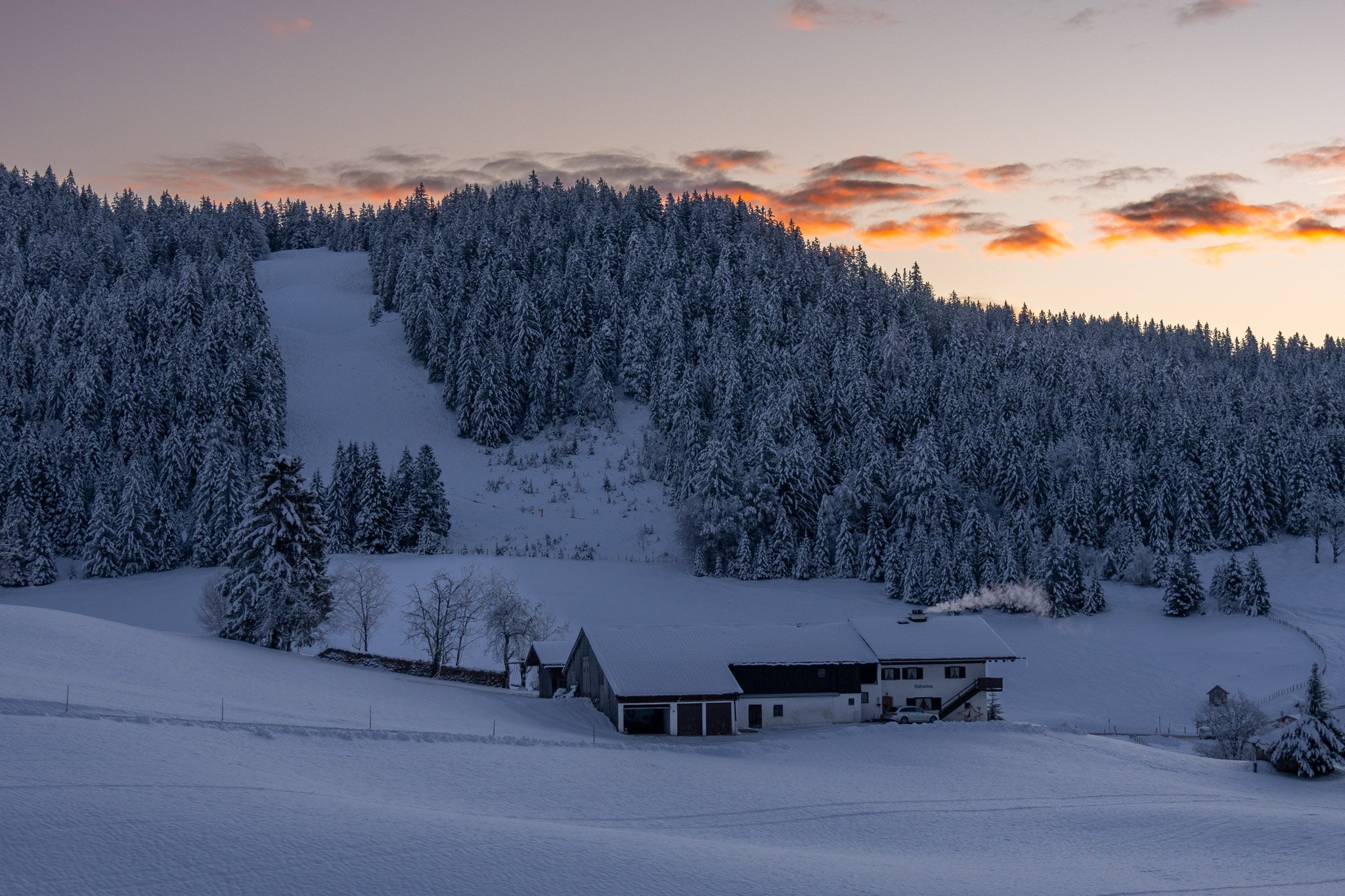 Snow-covered landscape with a small farm, surrounded by a dense forest of snow-laden trees, at sunset.