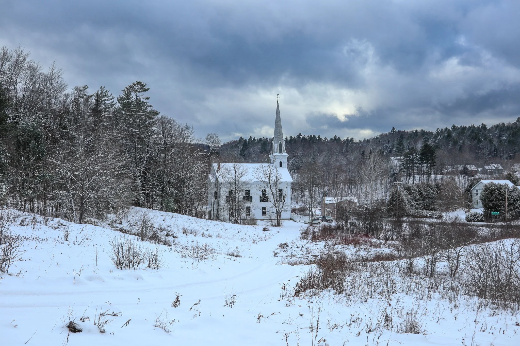 A white church with a tall steeple in a snowy landscape with bare trees and a cloudy sky.