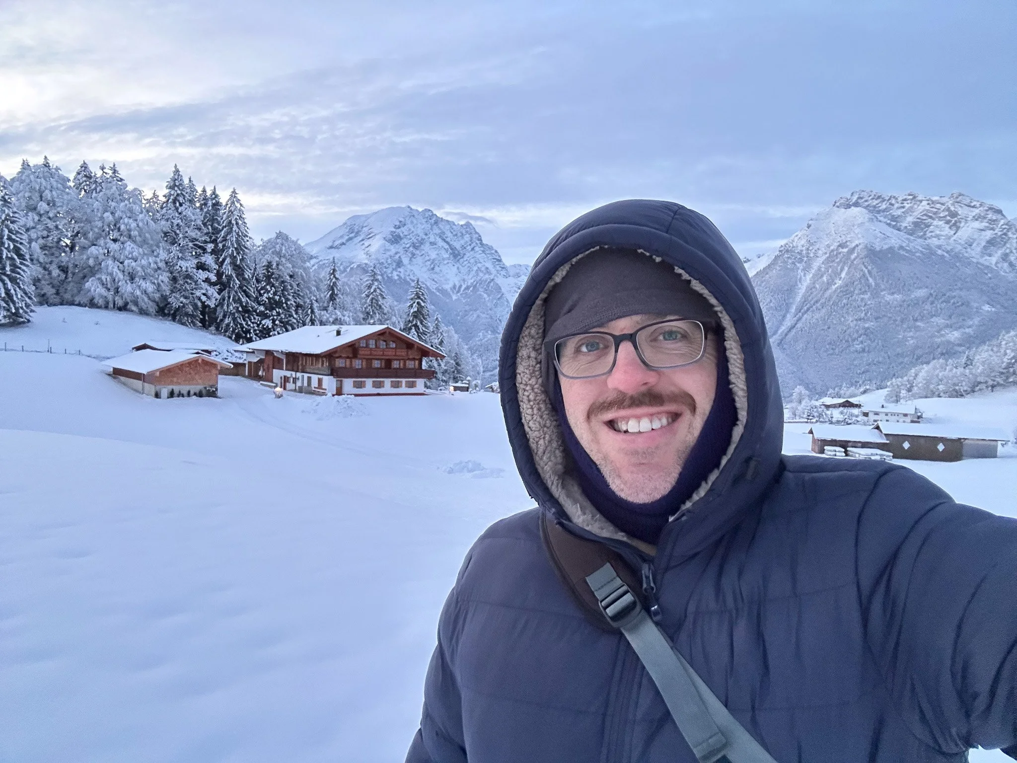 A man in winter clothing, including a navy jacket, gray hat, and glasses, takes a selfie in a snowy mountain landscape with wooden houses, snow-covered trees, and tall snow-capped mountains in the background.