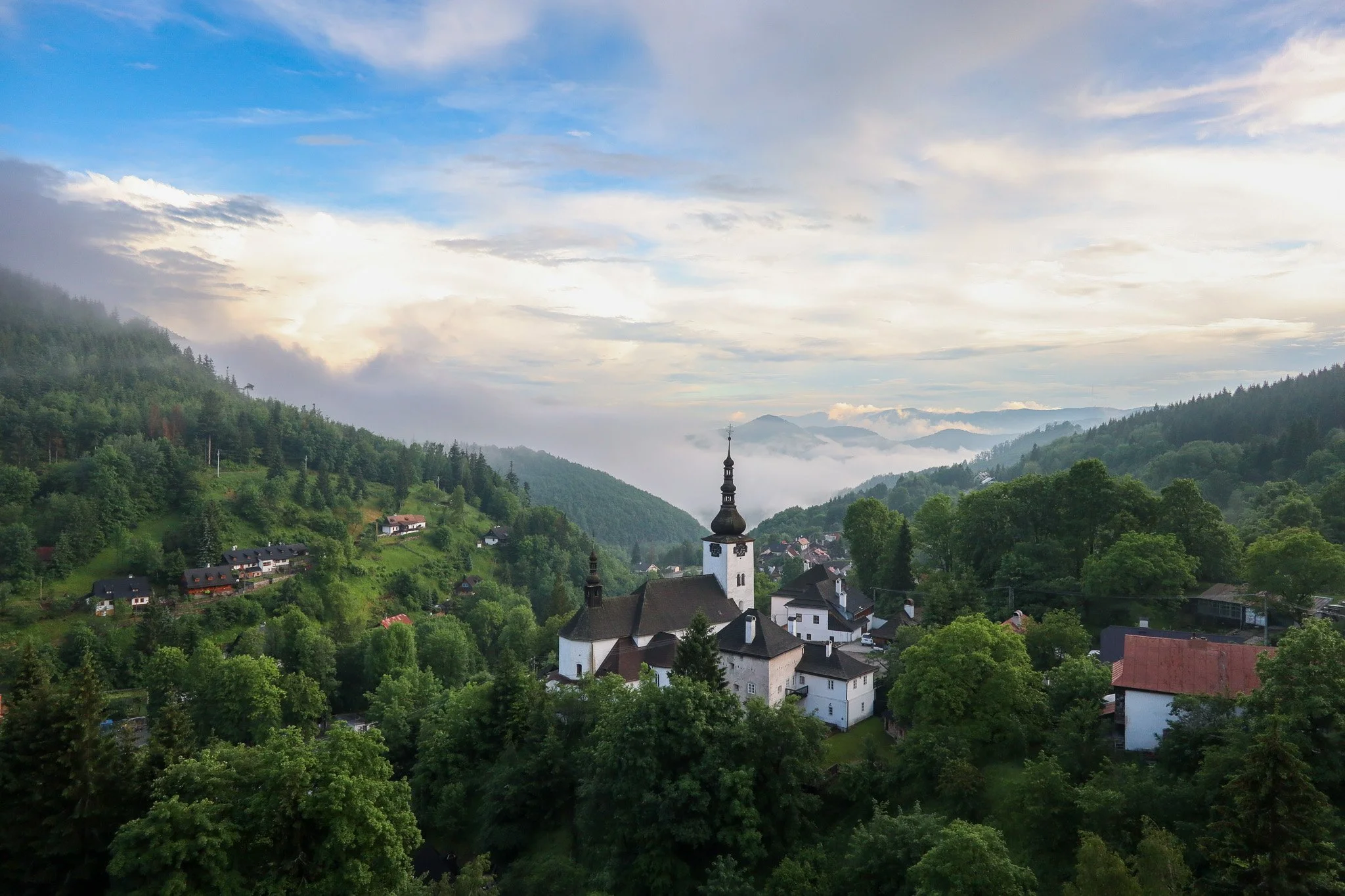 A scenic view of a small village with white buildings and a church with a tall steeple, surrounded by lush green hills and trees. In the background, mountains are visible through a misty sky with clouds and patches of blue.
