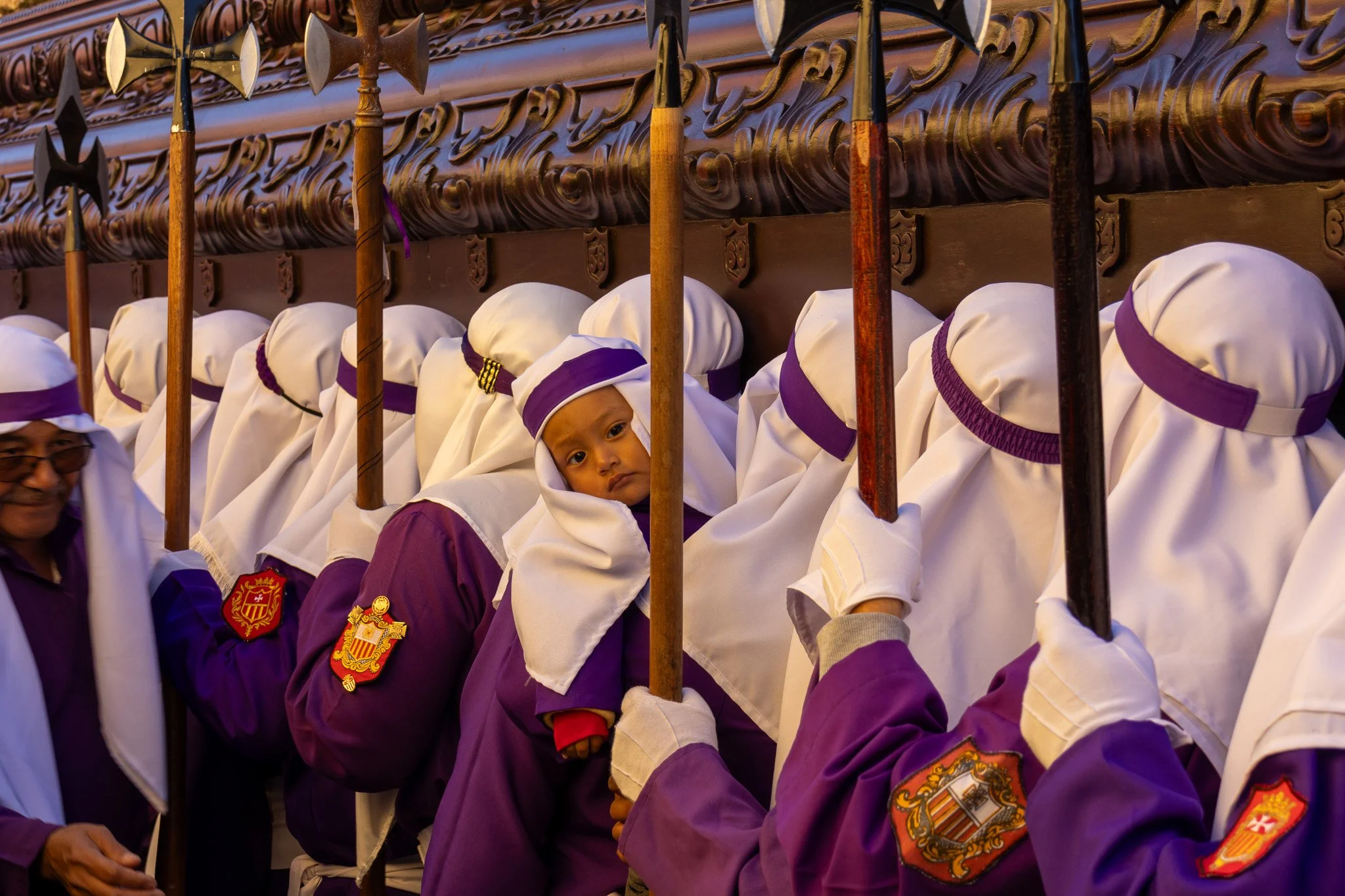 Young boy peers from behind a white hood with purple band, part of a group of people dressed in purple uniforms and white hoods, standing in front of a decorated wall with hanging wooden objects.