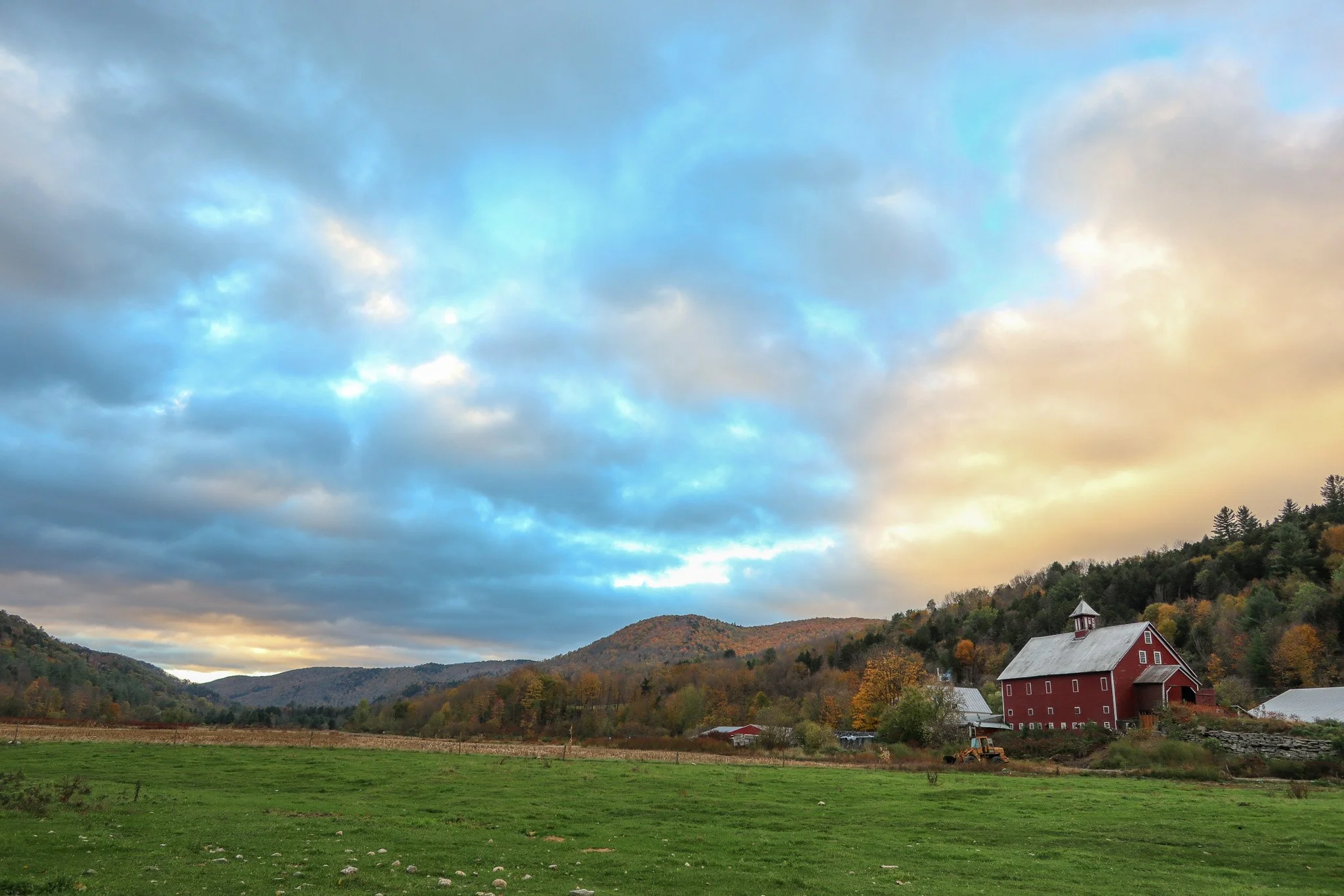 A red barn in a rural landscape with rolling hills and trees, under a partly cloudy sky during sunset.