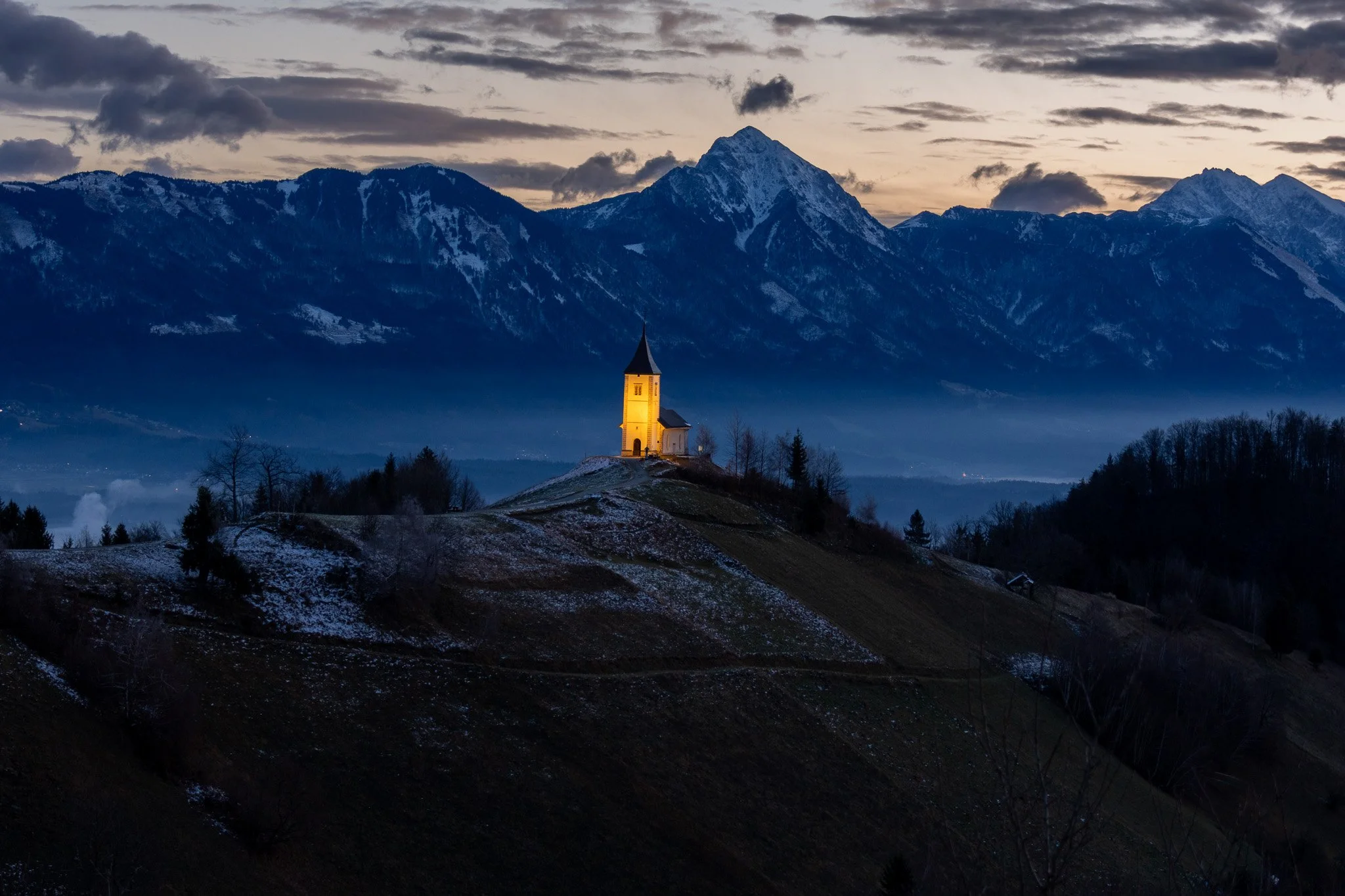A church with illuminated yellow light stands on a hill in a mountainous area during twilight or early evening, with snow on the ground and dark clouds in the sky.