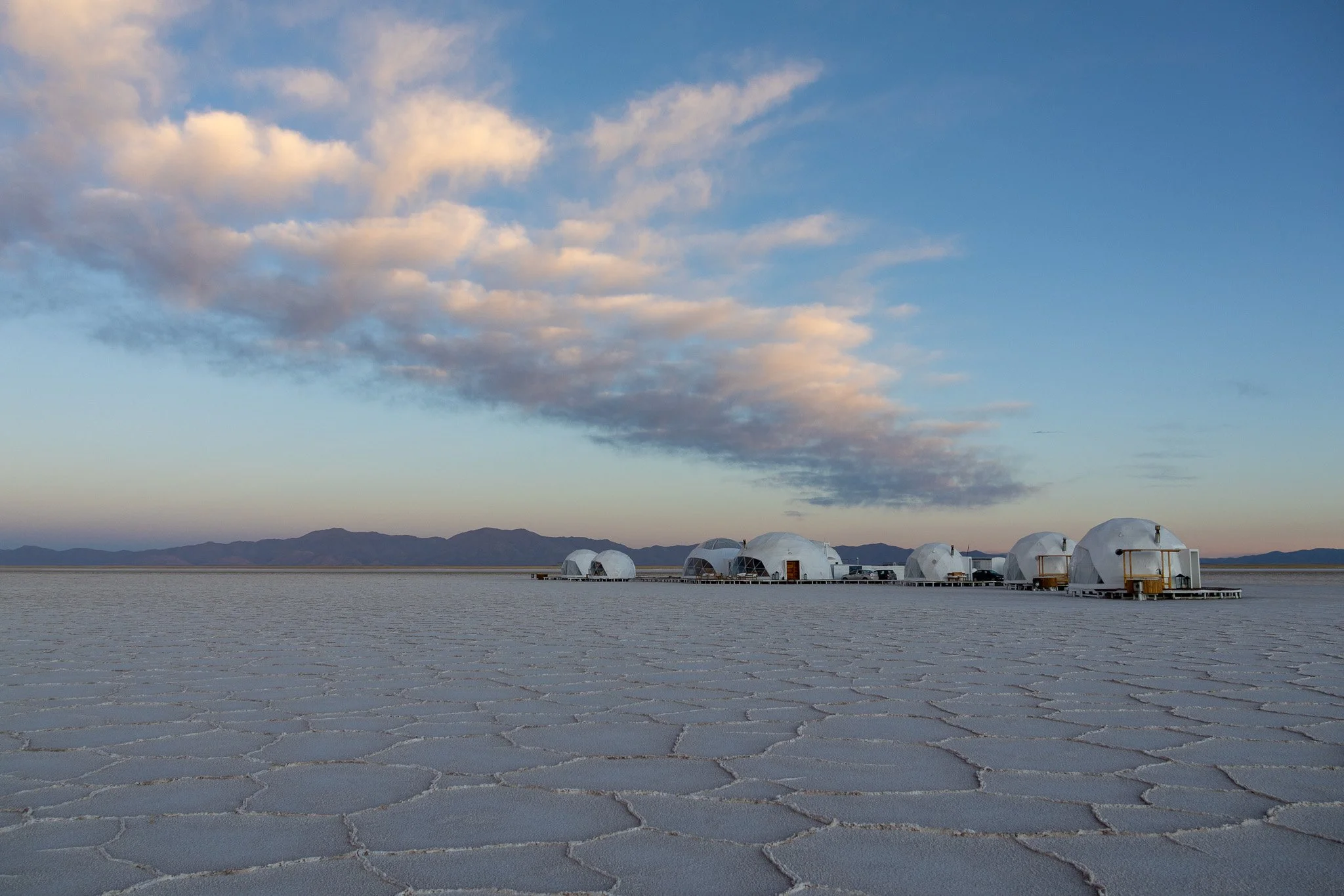 Cluster of futuristic white dome-shaped structures on salt flats with mountains in the distance and clouds in the sky.