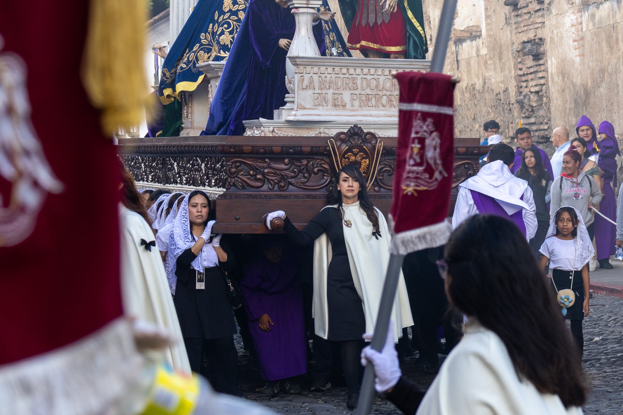 A religious procession with people carrying a large ornate float on their shoulders. Participants are dressed in traditional and ceremonial clothing, including white and purple garments. Some women wear lace veils, and others are dressed in black and