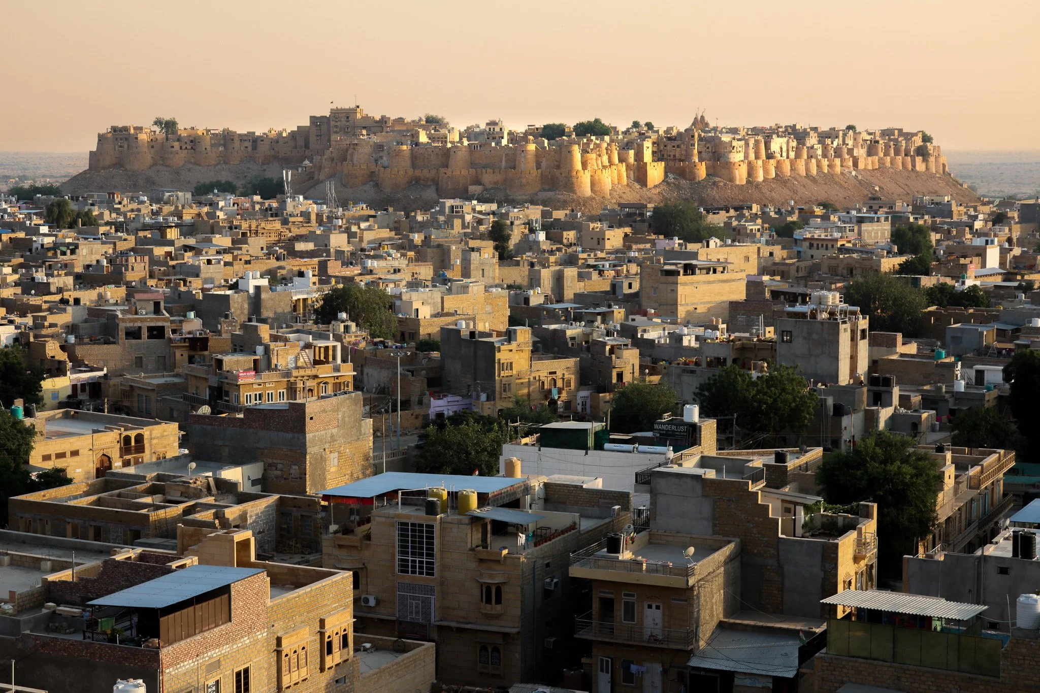 A cityscape with numerous beige and brown buildings, with a large fort on a hill in the background at sunset.