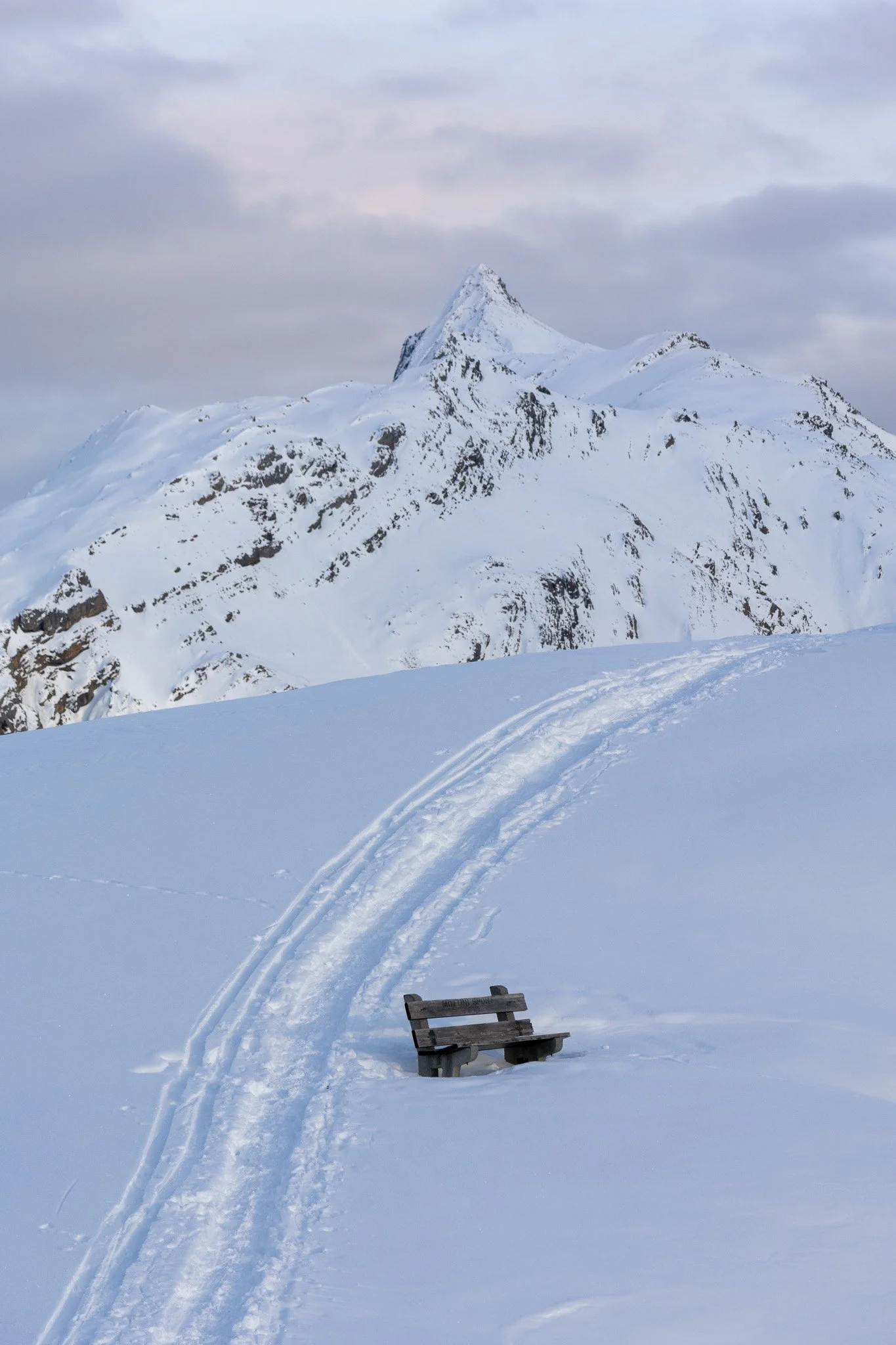 Snow-covered mountain landscape with ski tracks and a wooden bench in the foreground.