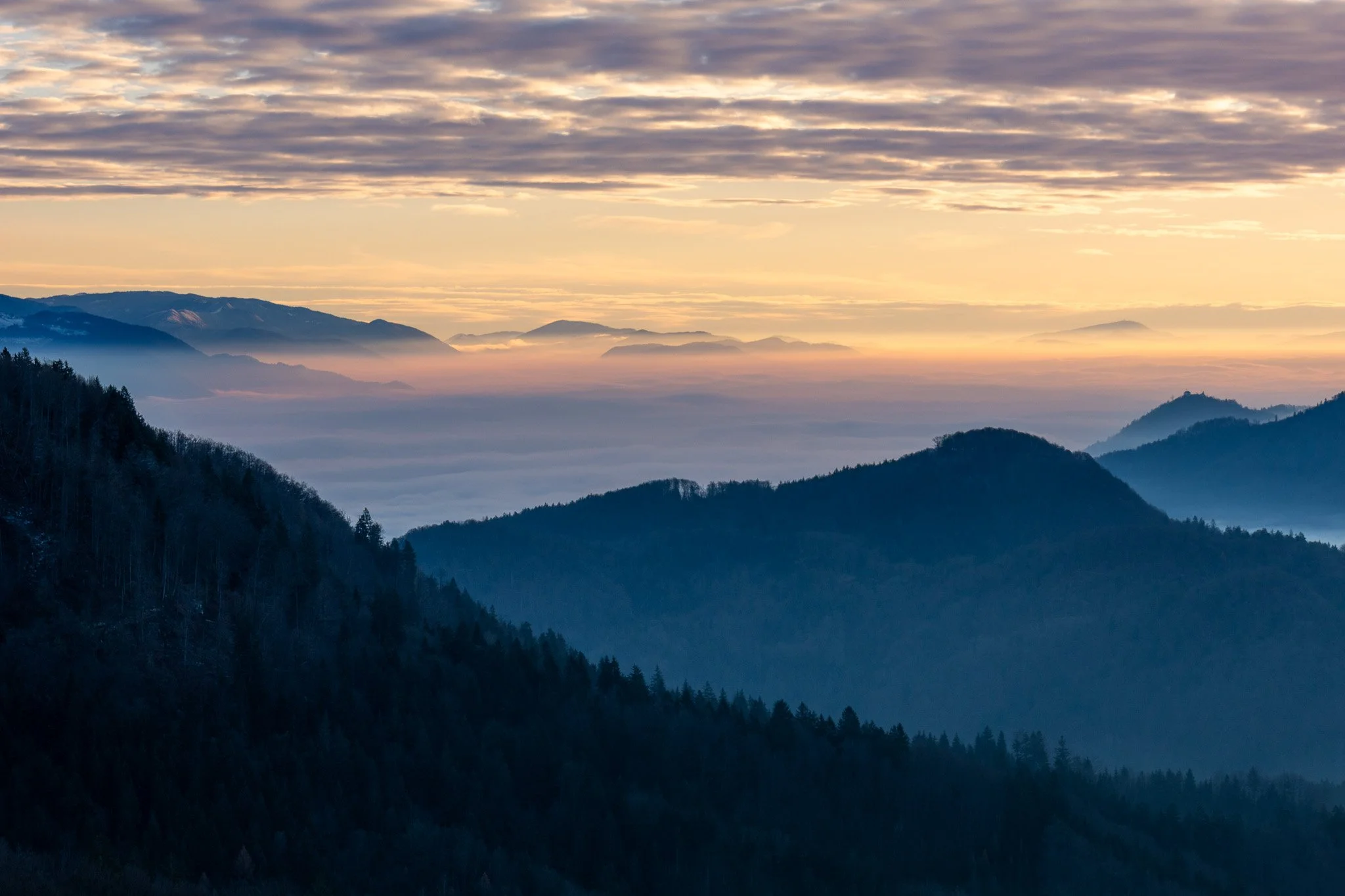 A scenic view of overlapping mountain ranges with a layer of clouds or fog, under a sky with scattered clouds during sunset or sunrise.