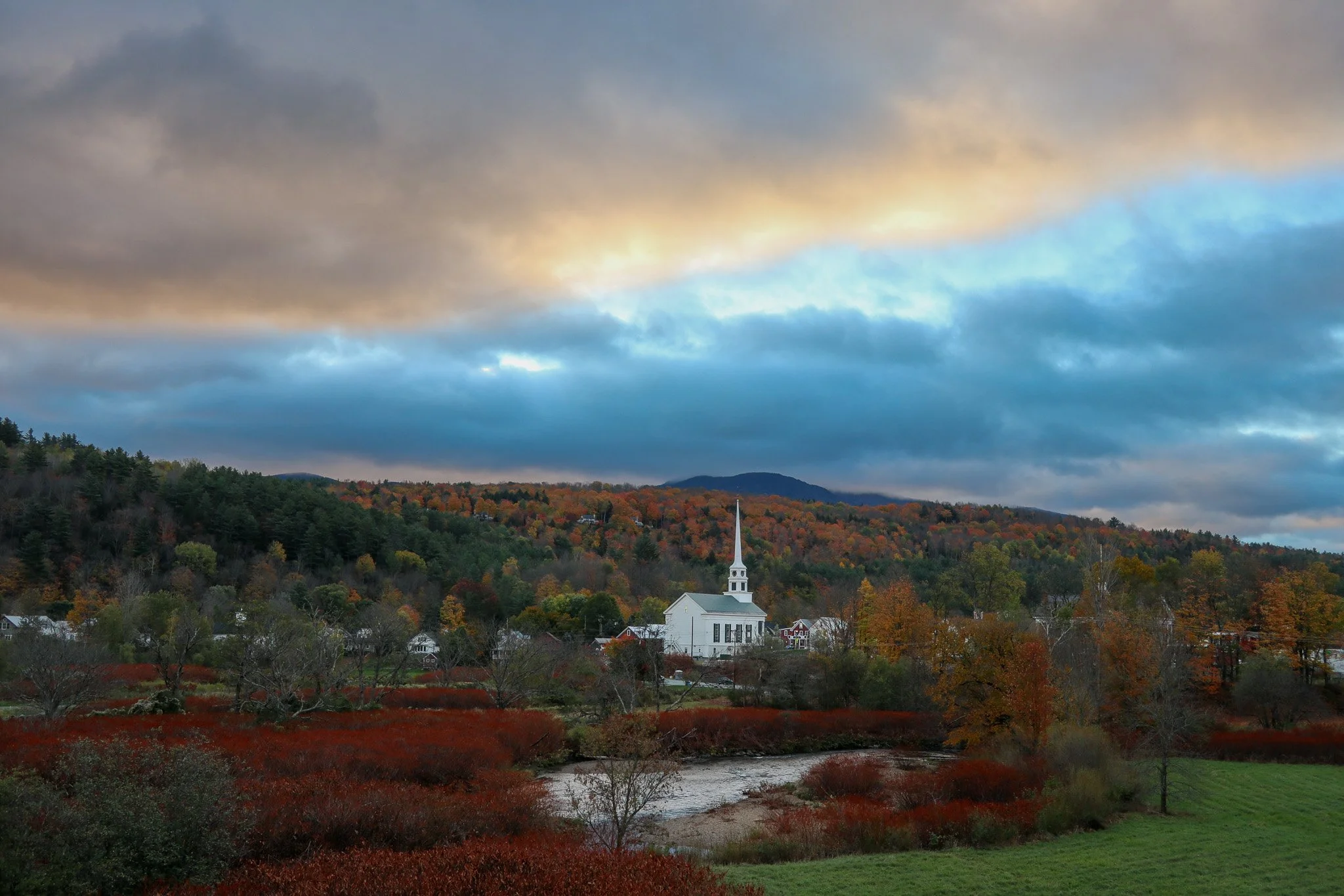A scenic view of a white church with a tall steeple situated in a rural area, surrounded by autumn-colored trees, a small river, and a green field, with mountains and a cloudy sky in the background.