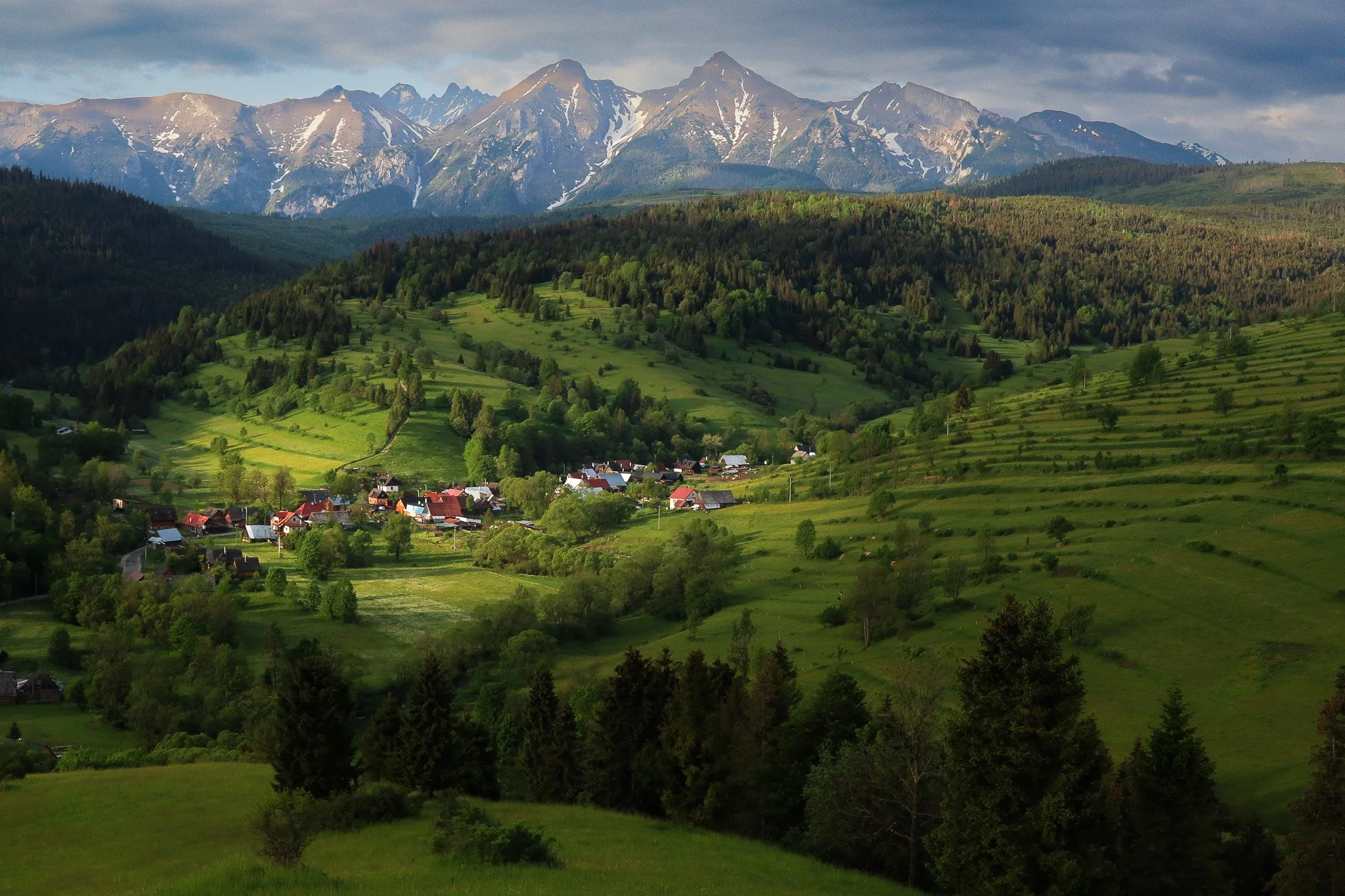 A scenic landscape of a small village surrounded by green hills and mountains with snow-capped peaks in the background.