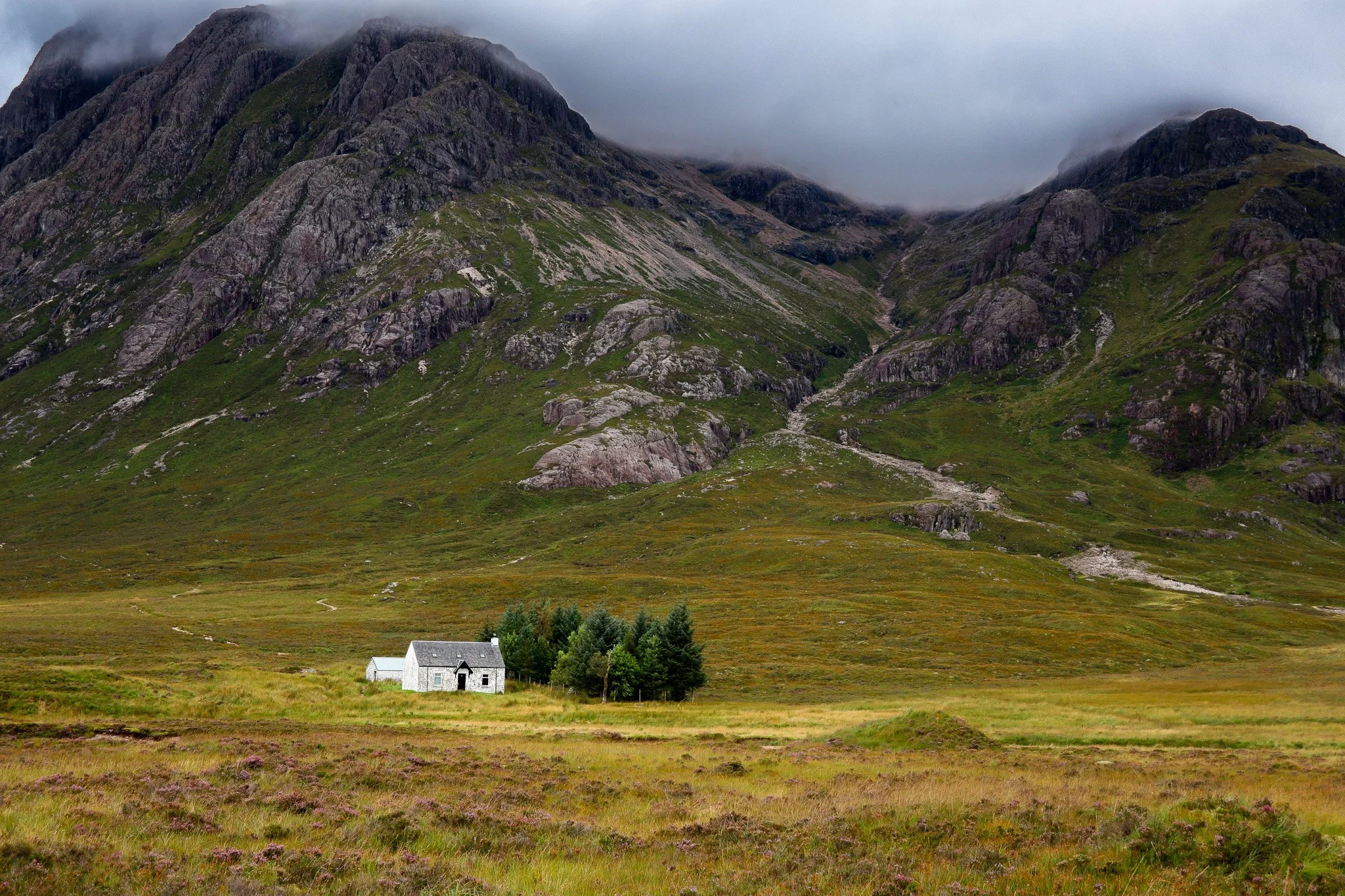 A small white house with a gray roof surrounded by green trees in a wide grassy valley, with steep rocky mountains partly covered by clouds in the background.