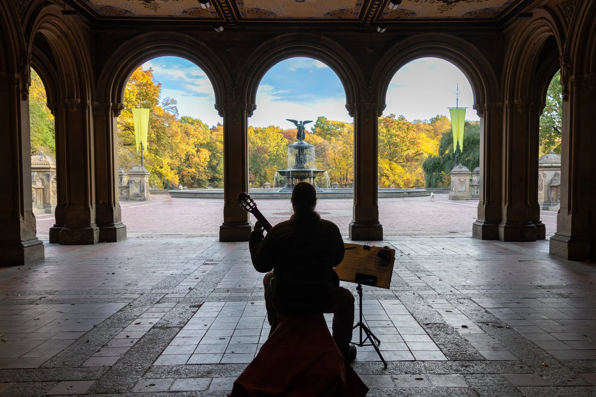 Silhouette of a person playing guitar and singing in a covered area with arches, looking out at a fountain with a statue and colorful autumn trees in the background.
