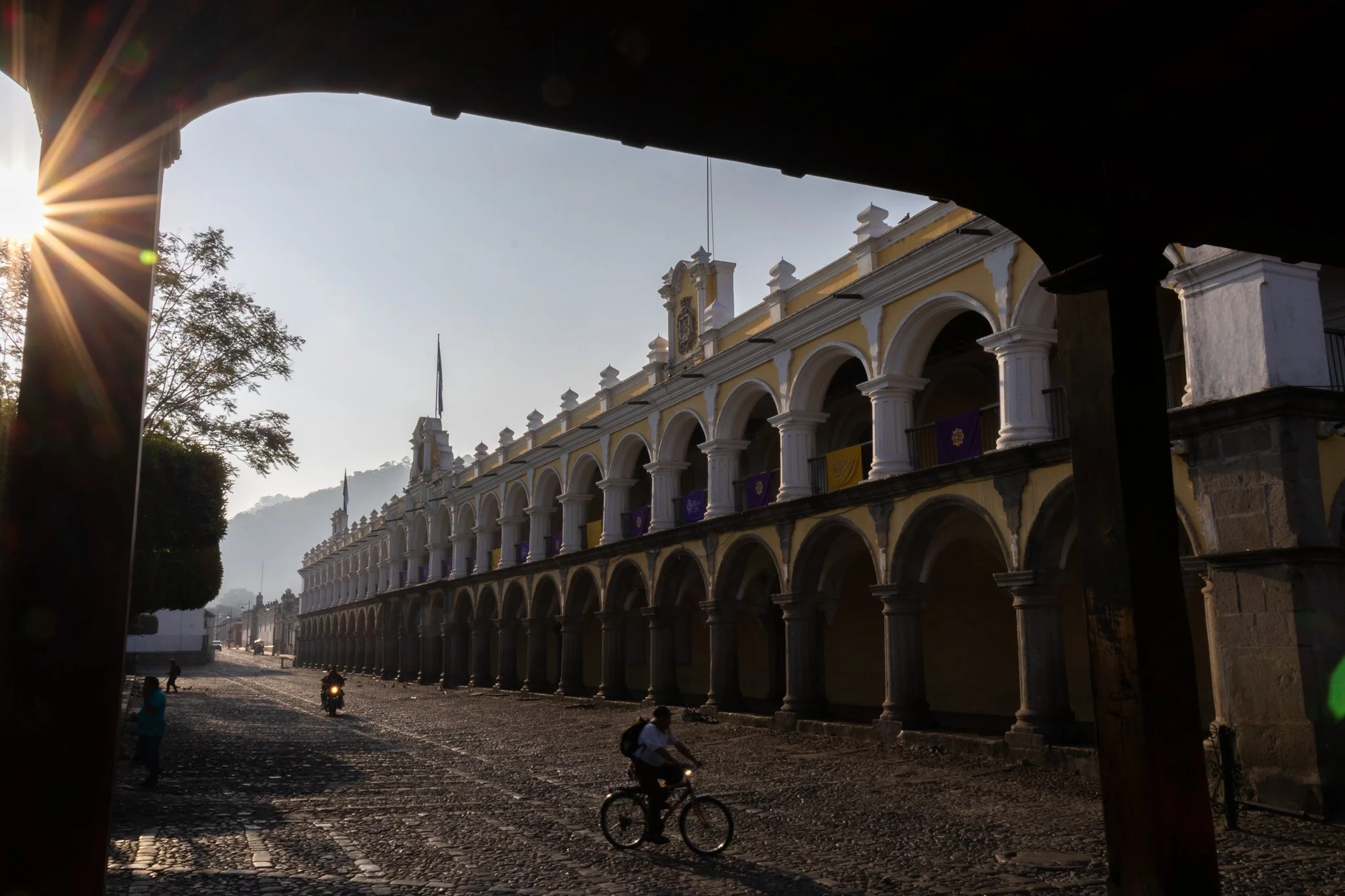 Historic yellow building with arches and white columns, viewed from under a wooden structure, with cobblestone street, bikers and pedestrians, in the early morning with sunlight