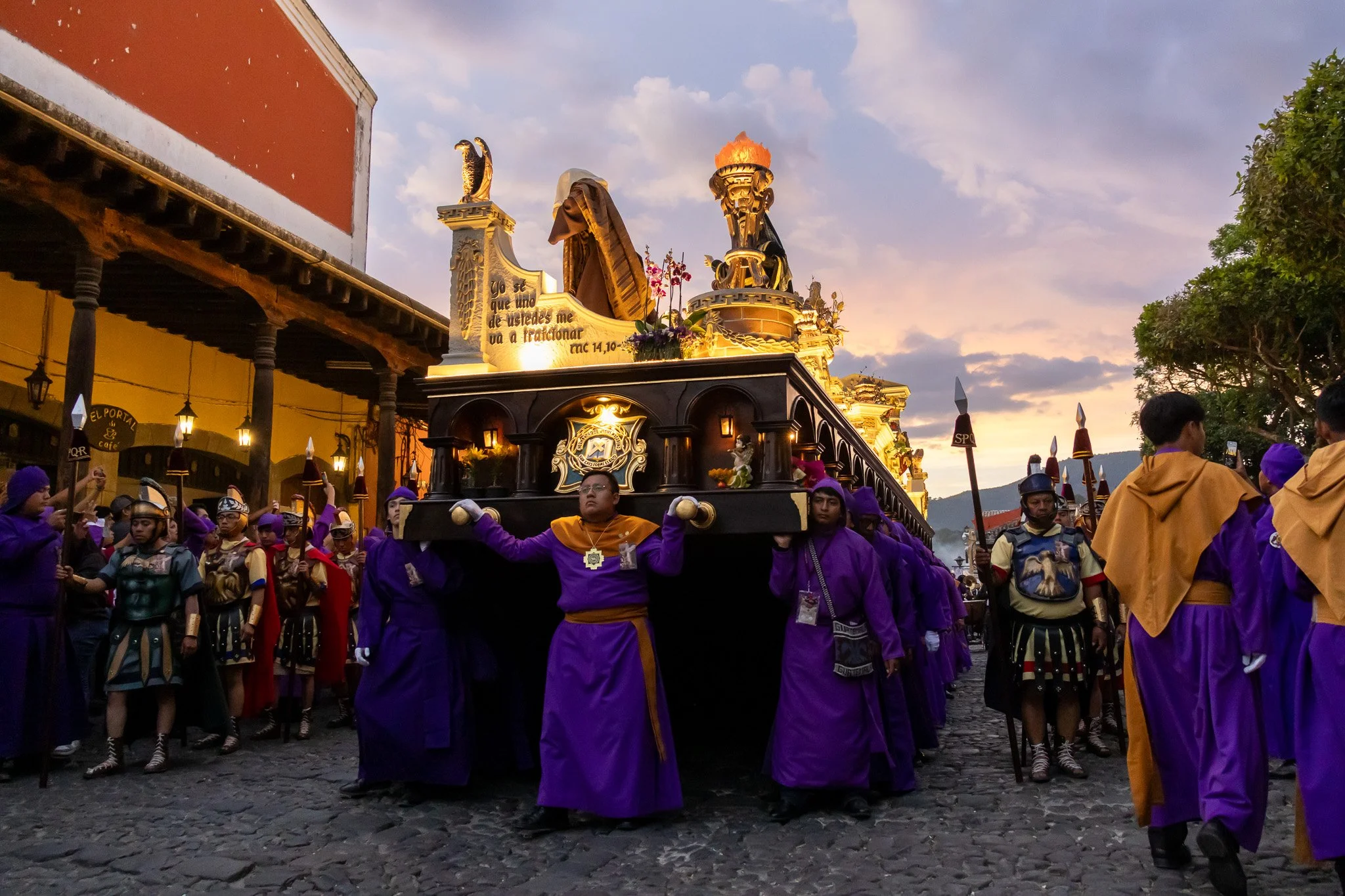 A procession with people dressed in colorful costumes carrying a religious float during a festival at sunset in a town square.