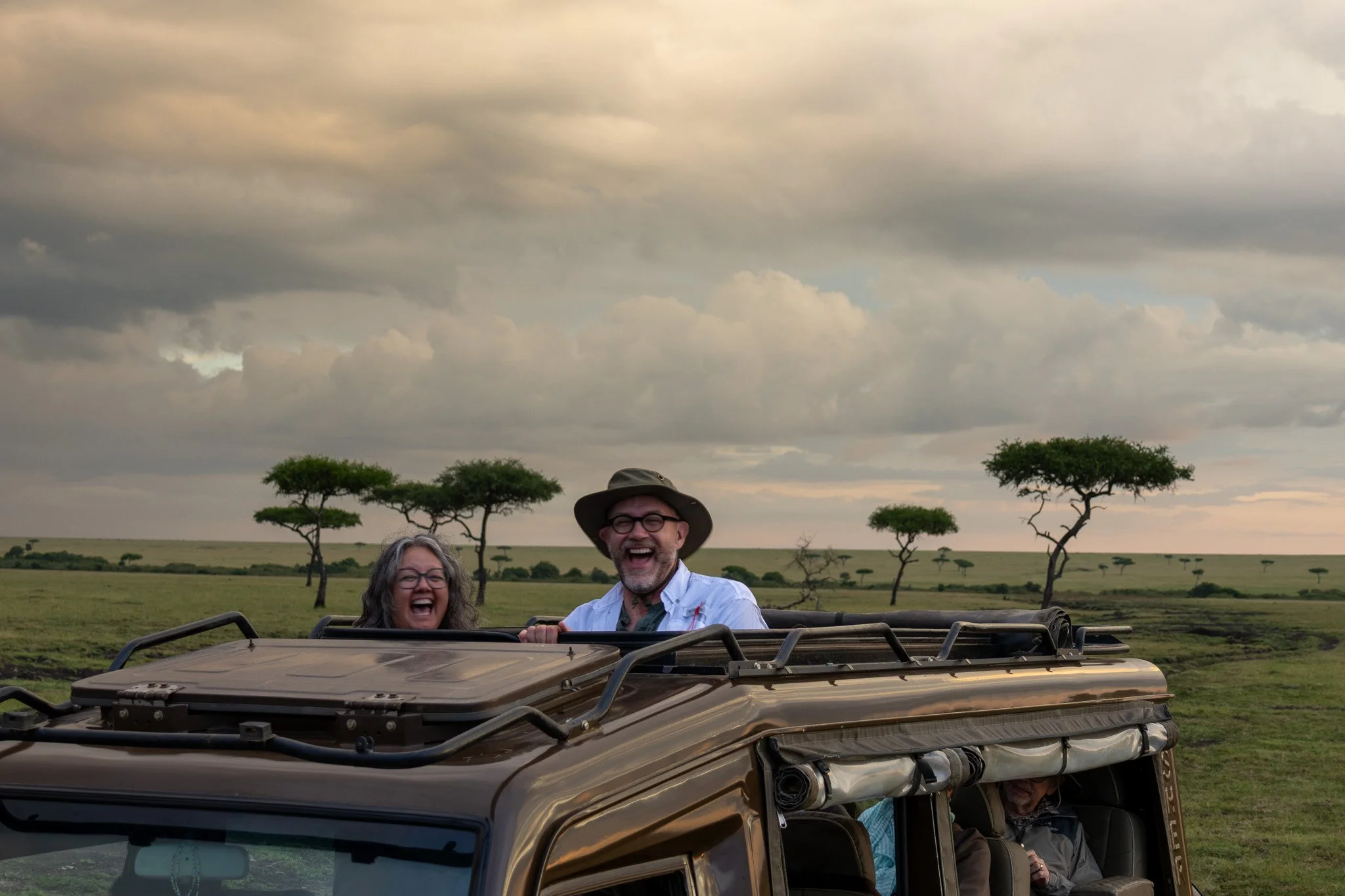 Two people on a safari vehicle enjoying a game drive in an open plain with acacia trees, under a cloudy sky.
