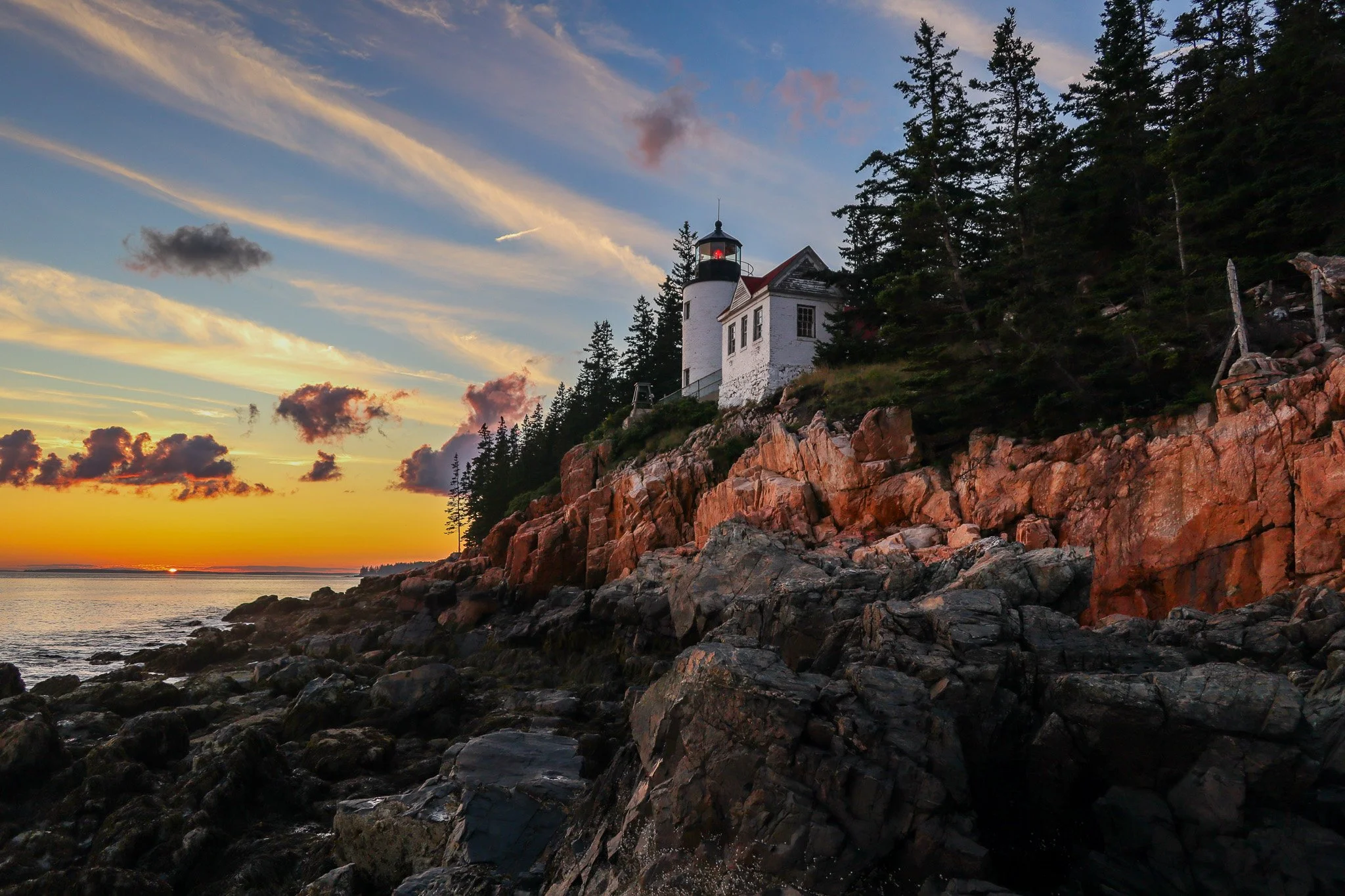 A white lighthouse on a rocky cliff at sunset, with green trees and a colorful sky with clouds in the background.