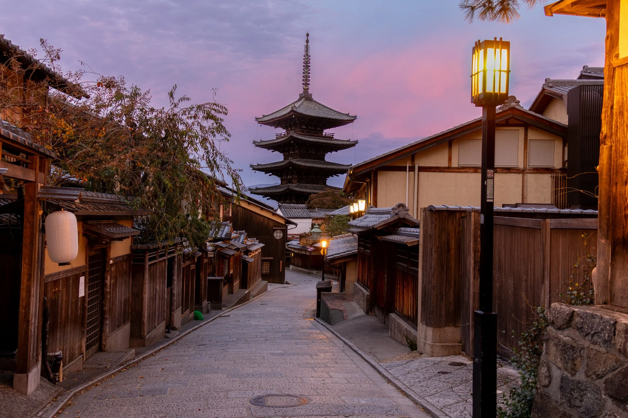 Traditional Japanese street at dusk with wooden houses, paper lanterns, and a five-story pagoda in the background, illuminated by streetlights and the fading pink and purple sky.