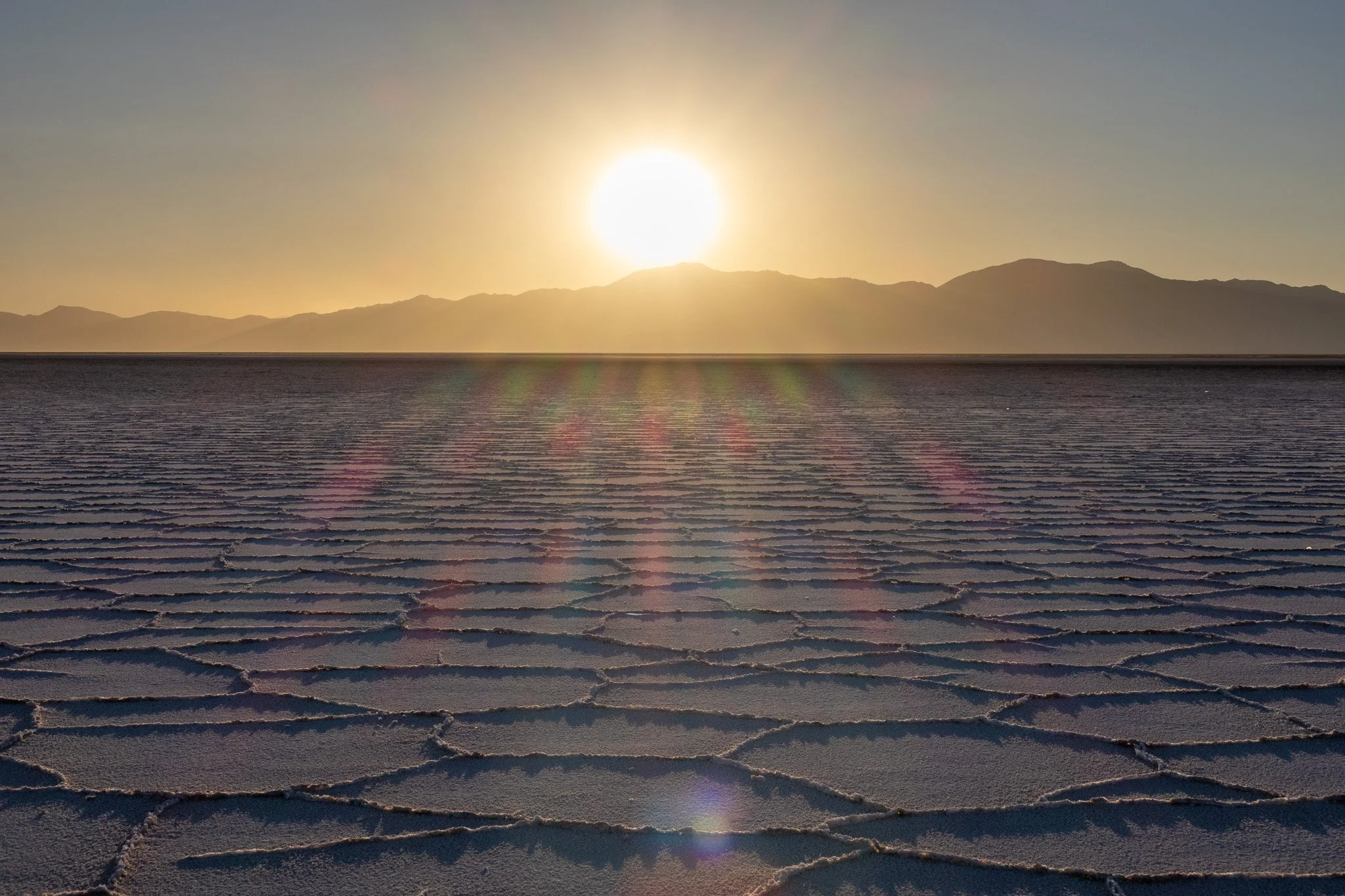 Sun setting over a salt flat with hexagonal salt formations and mountains in the background.
