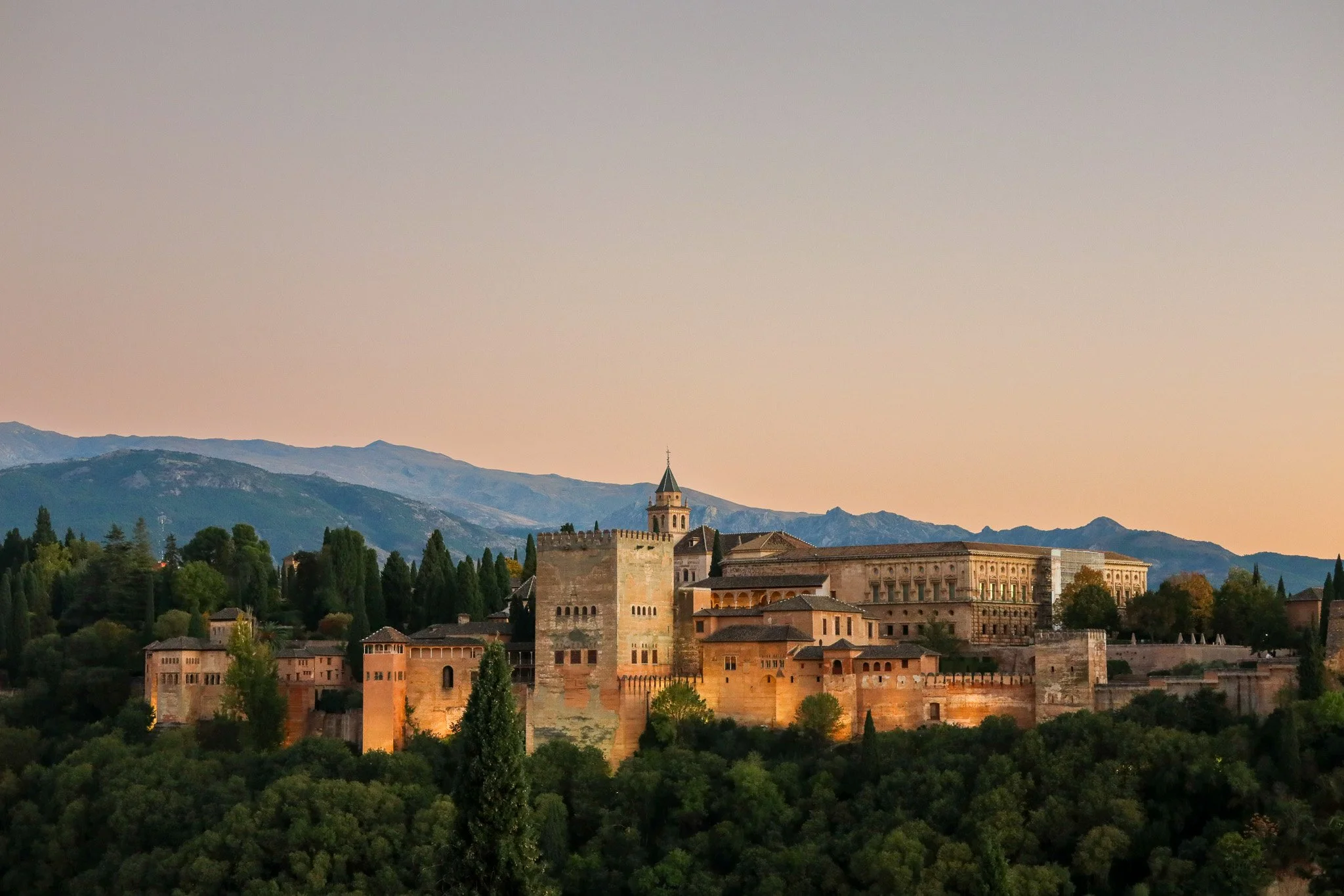 Sunset over the Alhambra Palace in Granada, Spain, with mountains in the background and lush green trees in the foreground.