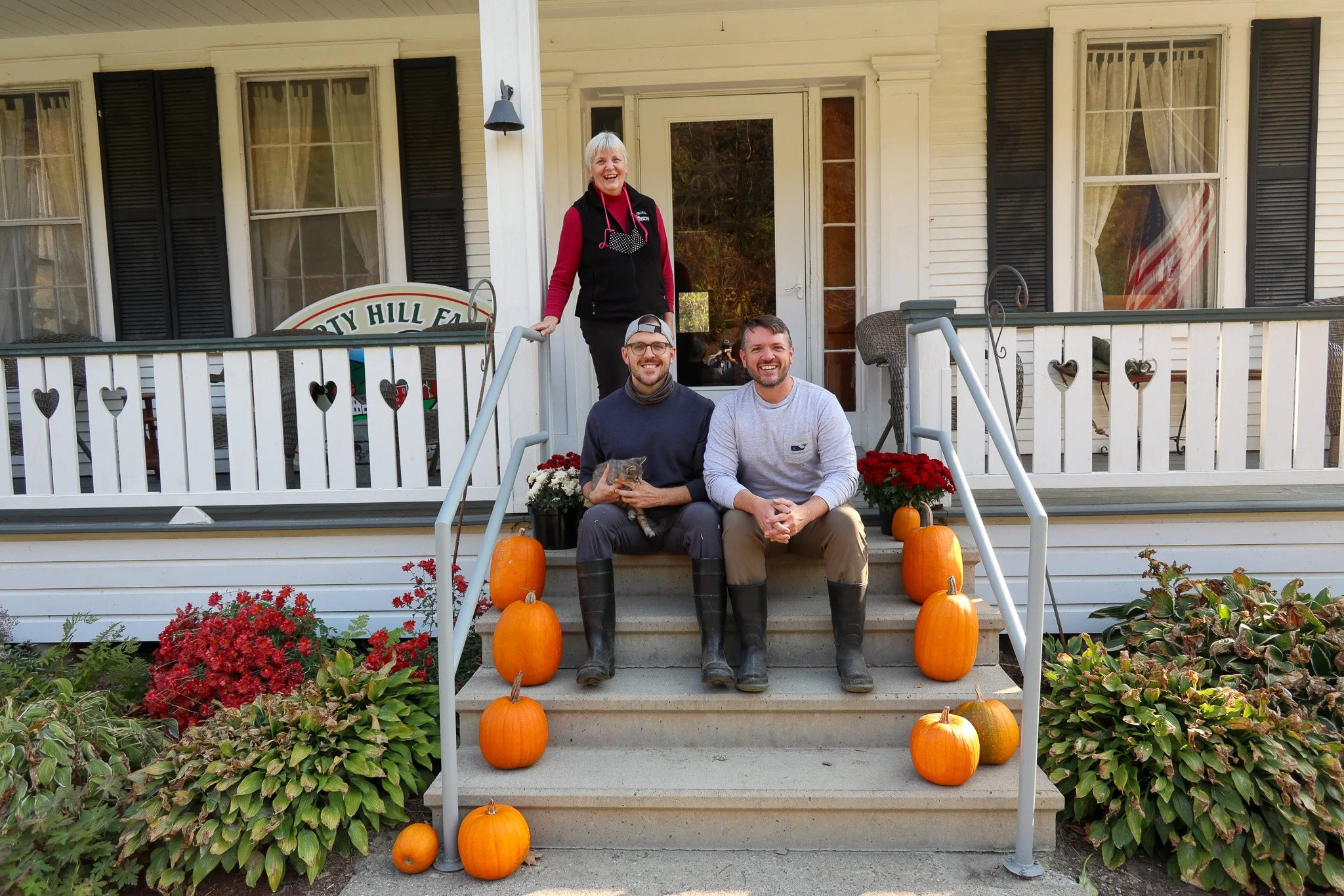 A group of four people sitting and standing on the front steps of a house decorated with pumpkins and flowers for fall, with two men sitting on the steps, a woman standing behind them, and another woman standing on the porch.
